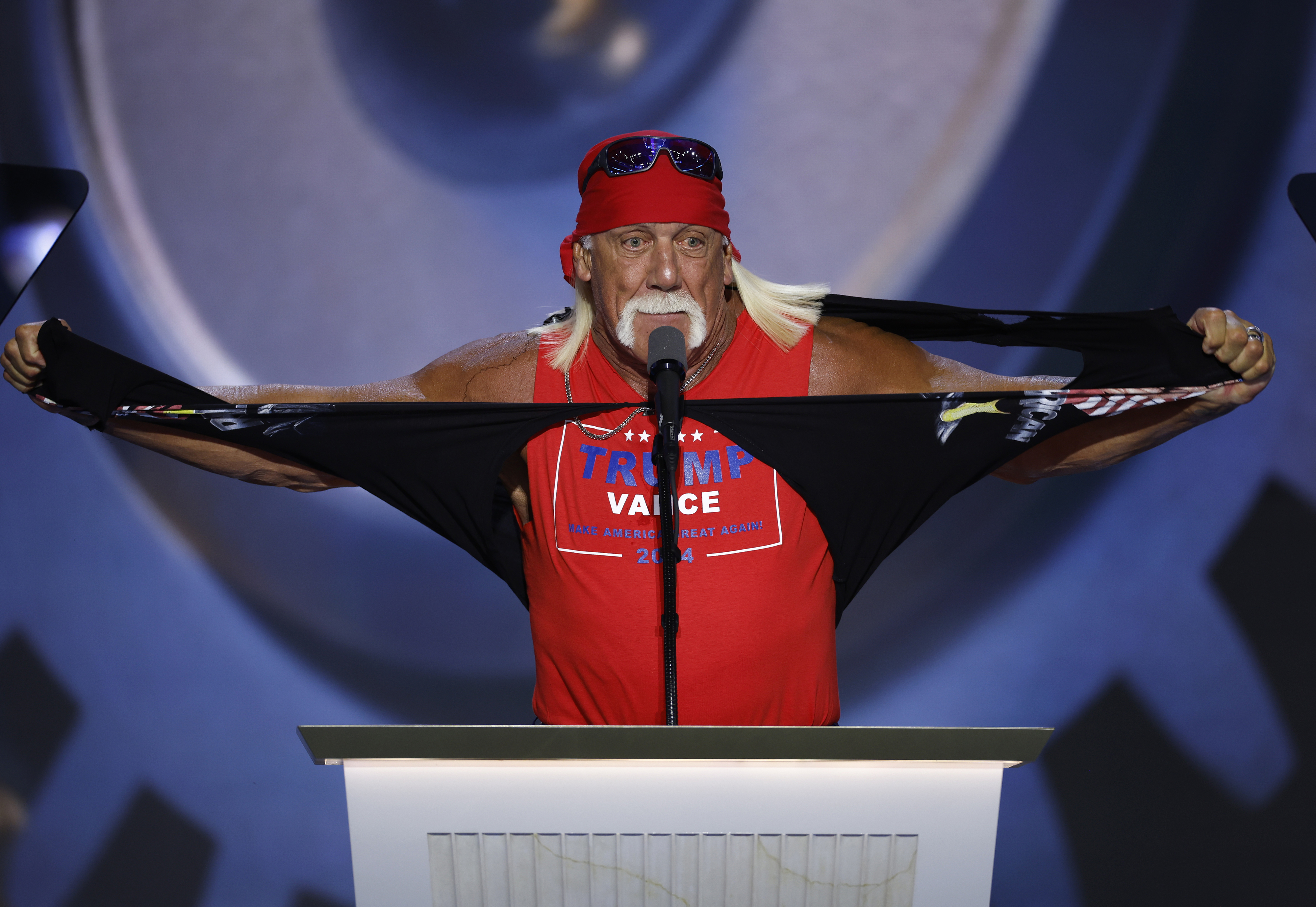 Hulk Hogan tearing a shirt while standing at a podium, wearing a red bandana, sunglasses, and a sleeveless &quot;Trump Pence&quot; shirt