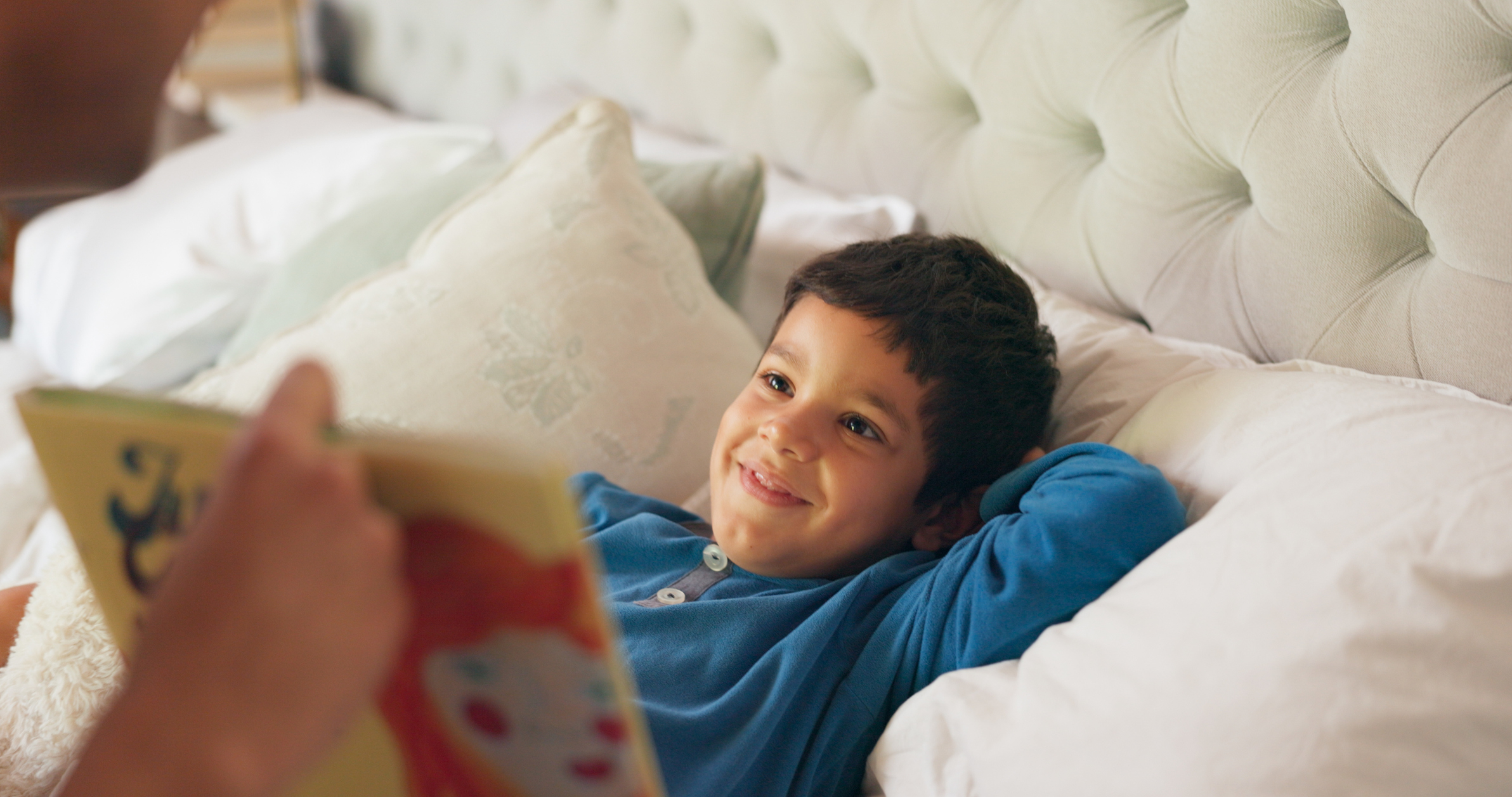 A young boy, lying in bed and smiling while looking at an adult who is holding a book with an illustration of a red-haired character on the cover