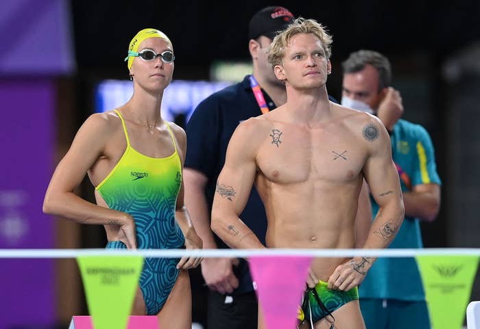 Swimming athletes Cody Simpson and Emma McKeon stand poolside at a sporting event. Emma wears a swimsuit and goggles, while Cody is in swim trunks