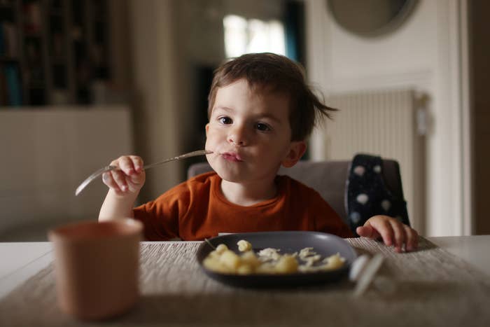 A young child with short hair sits at a table, eating food from a gray plate using a fork. A cup is placed in front of them