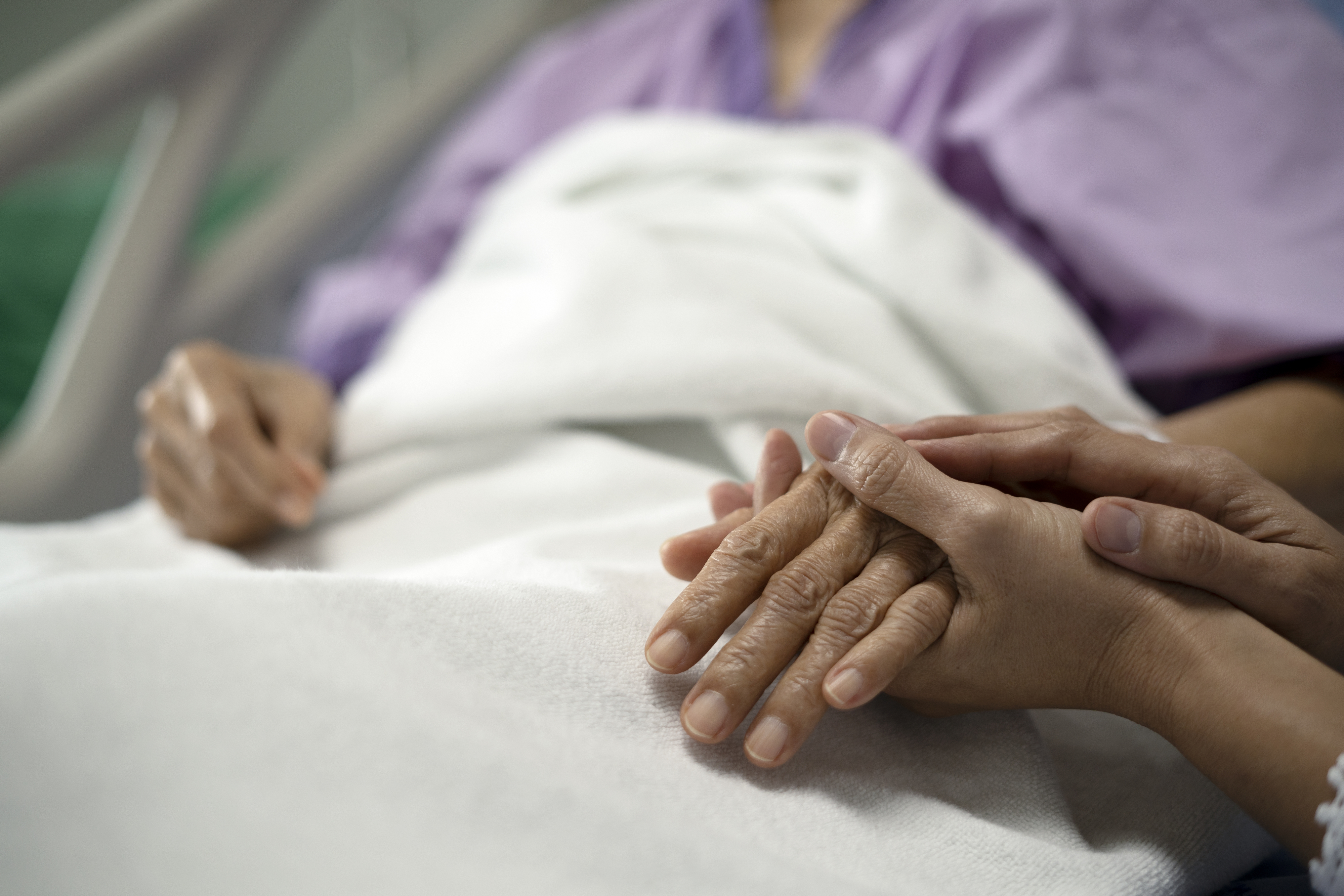 A person lies in a hospital bed with another person gently holding their hand, providing comfort and support
