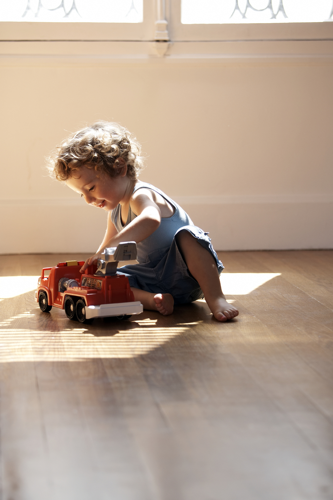 A young child with curly hair plays on the floor with a toy fire truck in a sunlit room