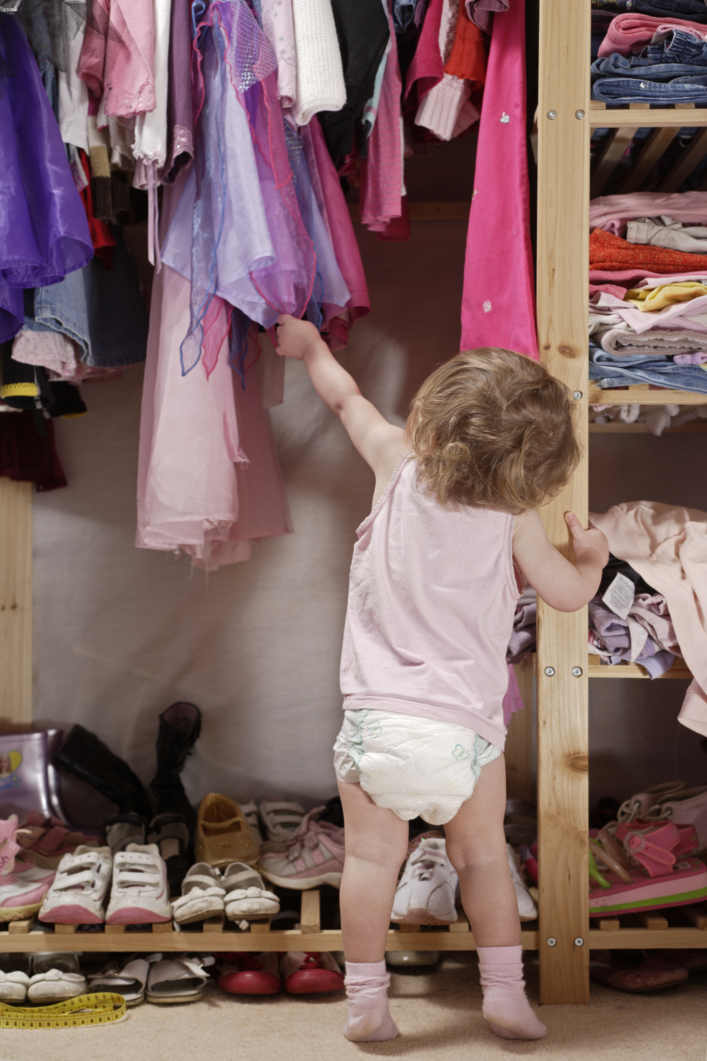 A toddler dressed in a diaper and tank top reaches for clothes on a high shelf in a closet filled with various clothing and shoes
