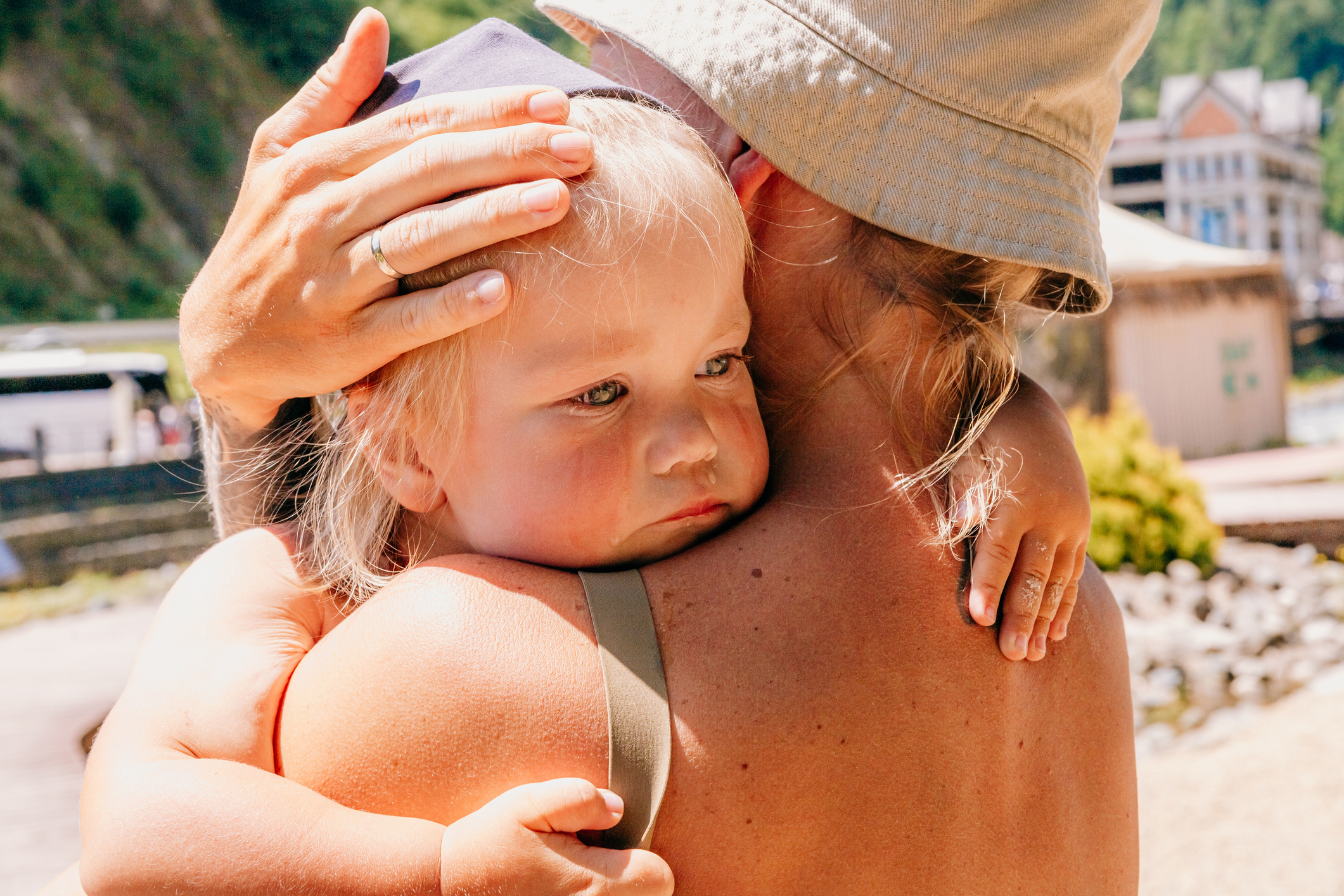 An adult wearing a hat holds a small, tired-looking child close, gently resting their hand on the child's head outdoors