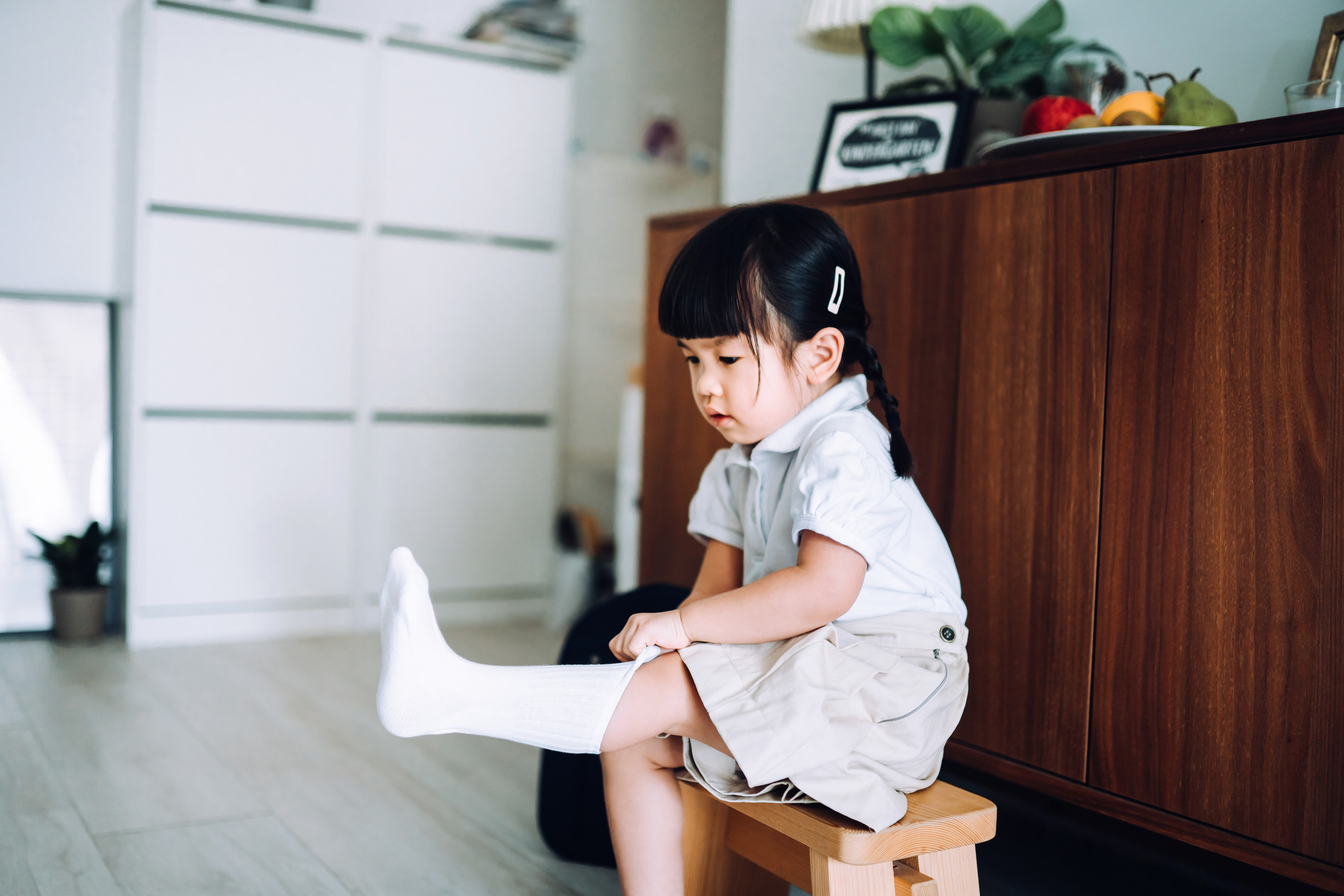 Young child with pigtails, seated on a stool, putting on white socks in a tidy room with a modern aesthetic