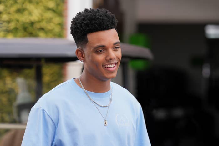 A young man, dressed in a casual blue shirt and silver necklace, smiles while standing outdoors