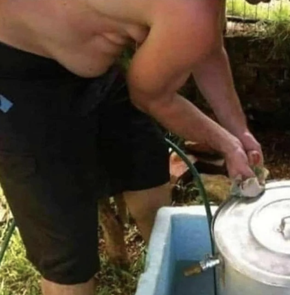 Person in shorts washing a large metal pot outdoors using a hose