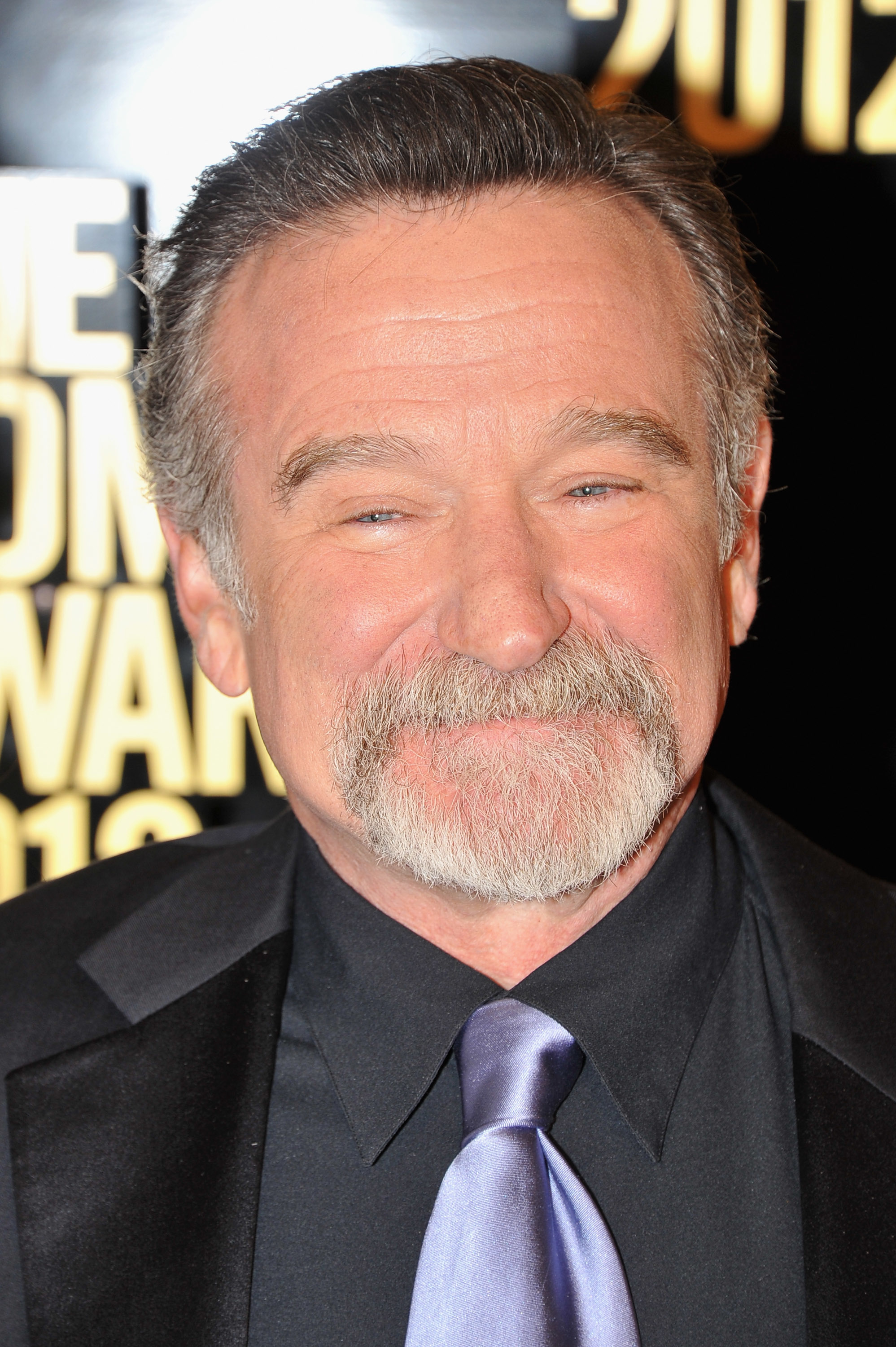 Robin Williams is smiling while wearing a suit and tie at an event. The background includes part of a sign with the year "2012"