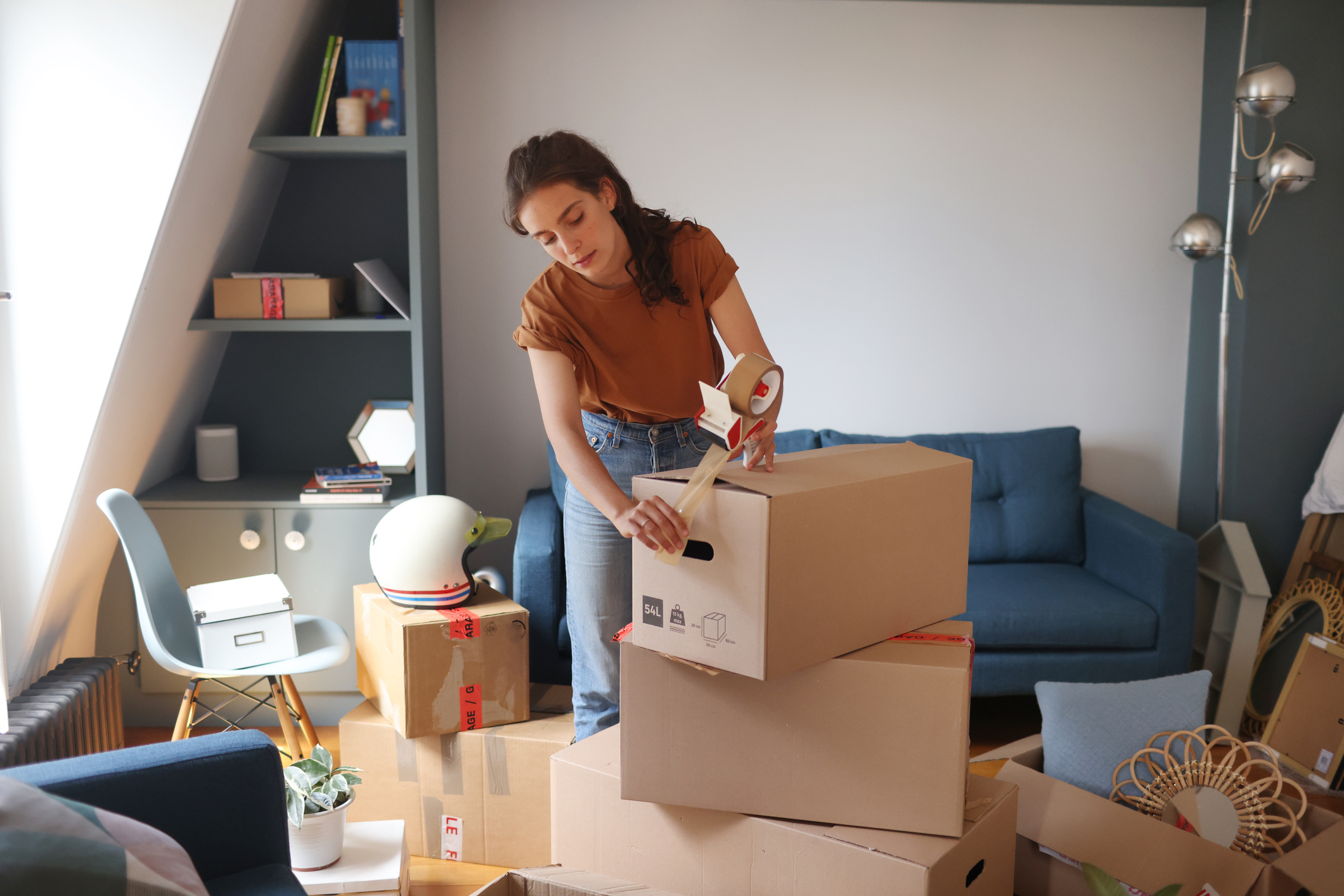 A woman is taping boxes in a small apartment with packed items, suggesting she is moving. She is wearing casual clothing and a focused expression