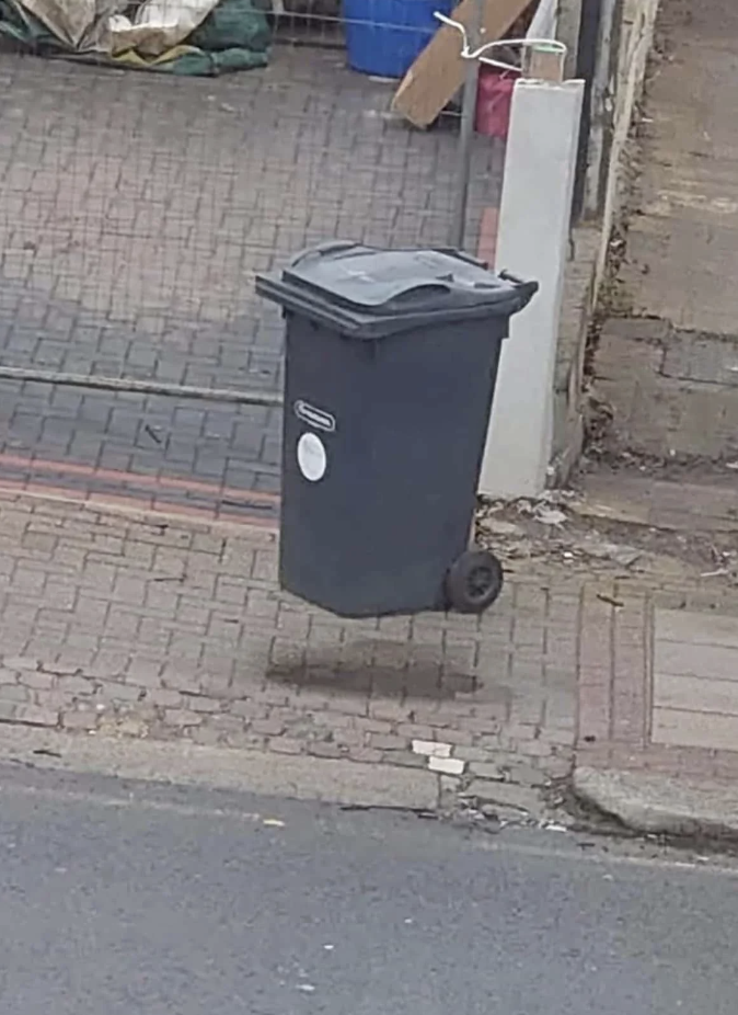 A wheelie trash bin appears to be floating off the ground on a brick-paved sidewalk near a street and garden area