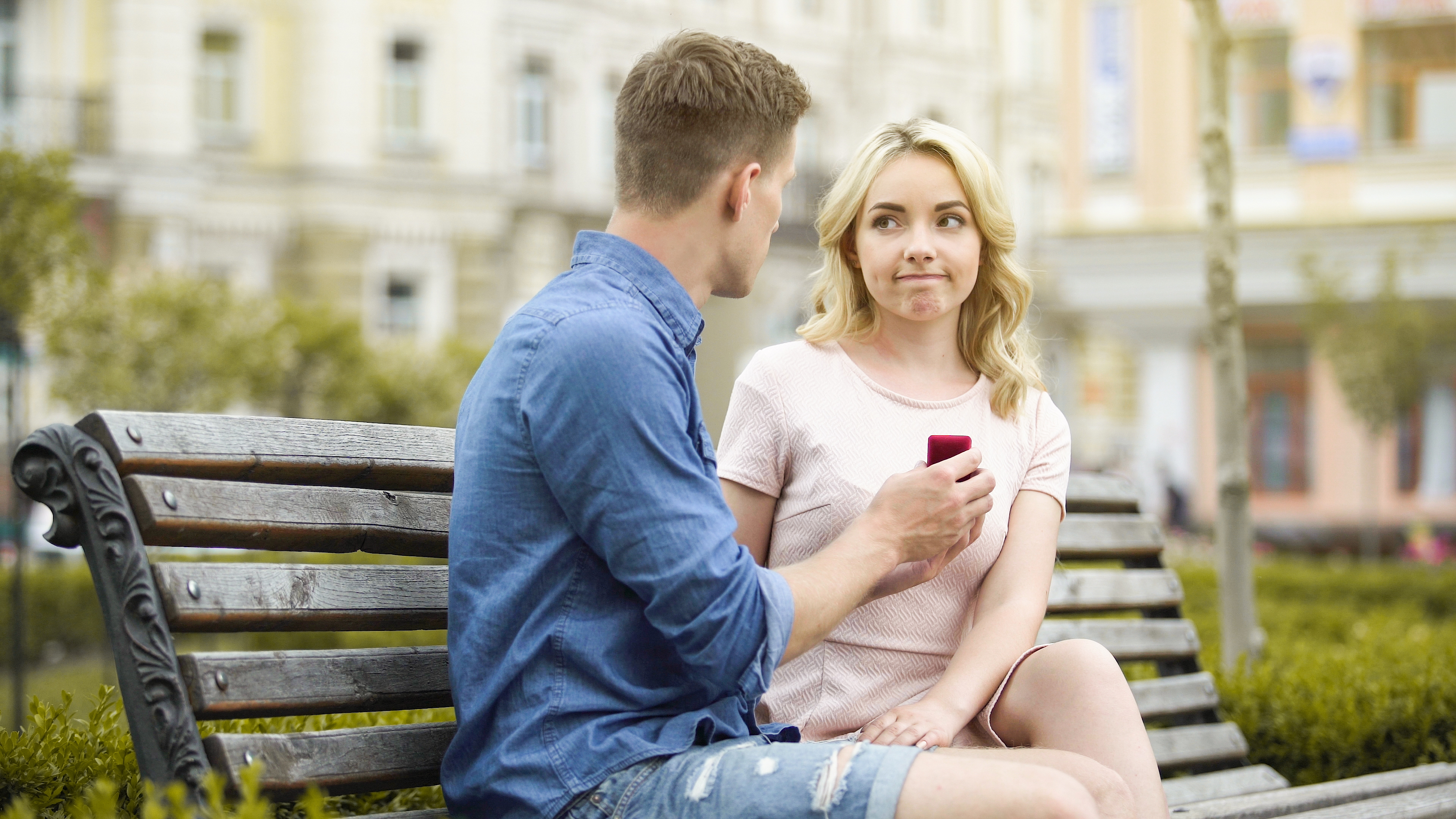 A man in a blue shirt and denim shorts proposing to a hesitant blonde woman on a park bench with buildings in the background