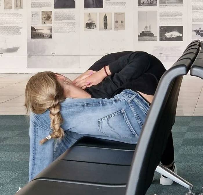 Two individuals stretching on waiting chairs, one with a braided hairstyle and casual denim, surrounded by historical photo displays in an airport or similar space