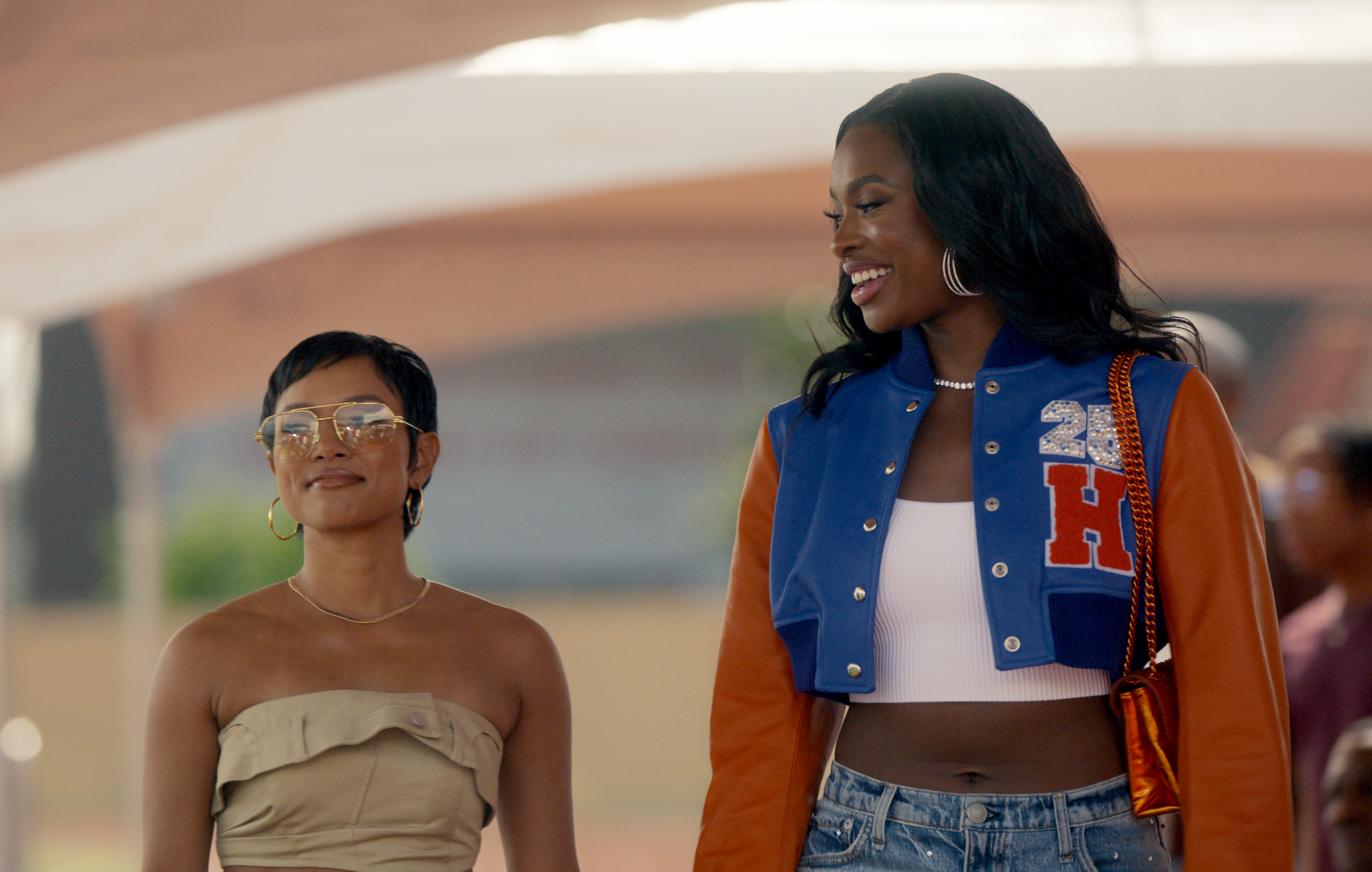Two women standing and smiling; one wears glasses and a strapless top, the other wears a varsity jacket with the letter &quot;H&quot; and a crop top. Names: Unknown