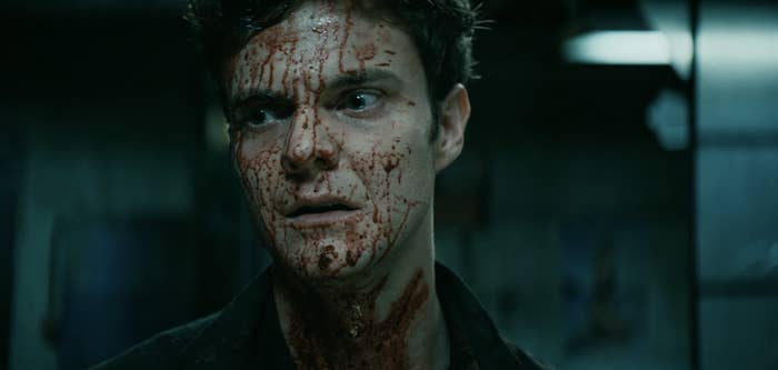 Jack Quaid with face and neck covered in blood, looking shocked in a dimly lit room. Scene from a movie or TV show