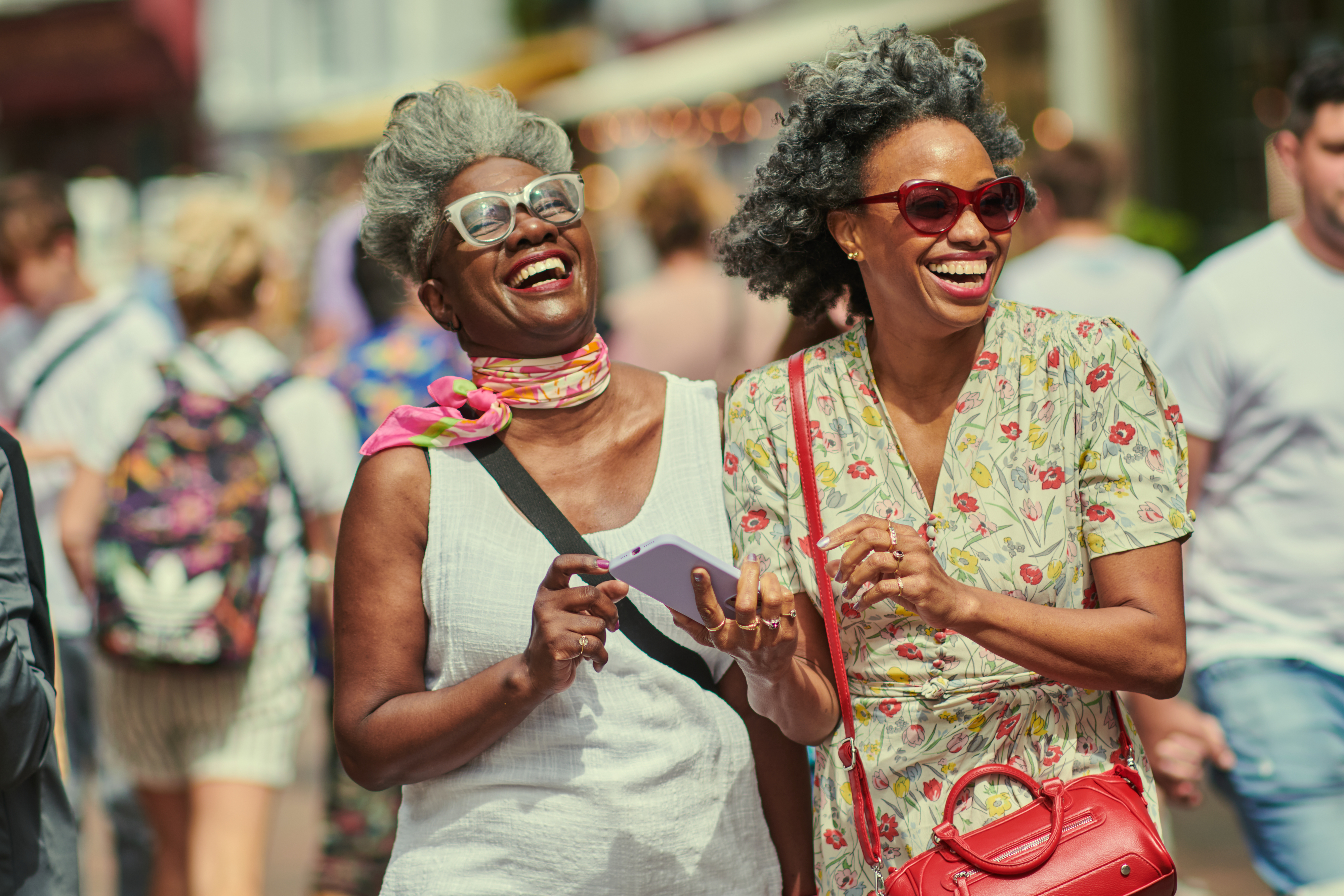 Two women, one in a white dress with a colorful scarf, and the other in a floral dress carrying a red purse, joyfully walking outdoors, smiling and laughing