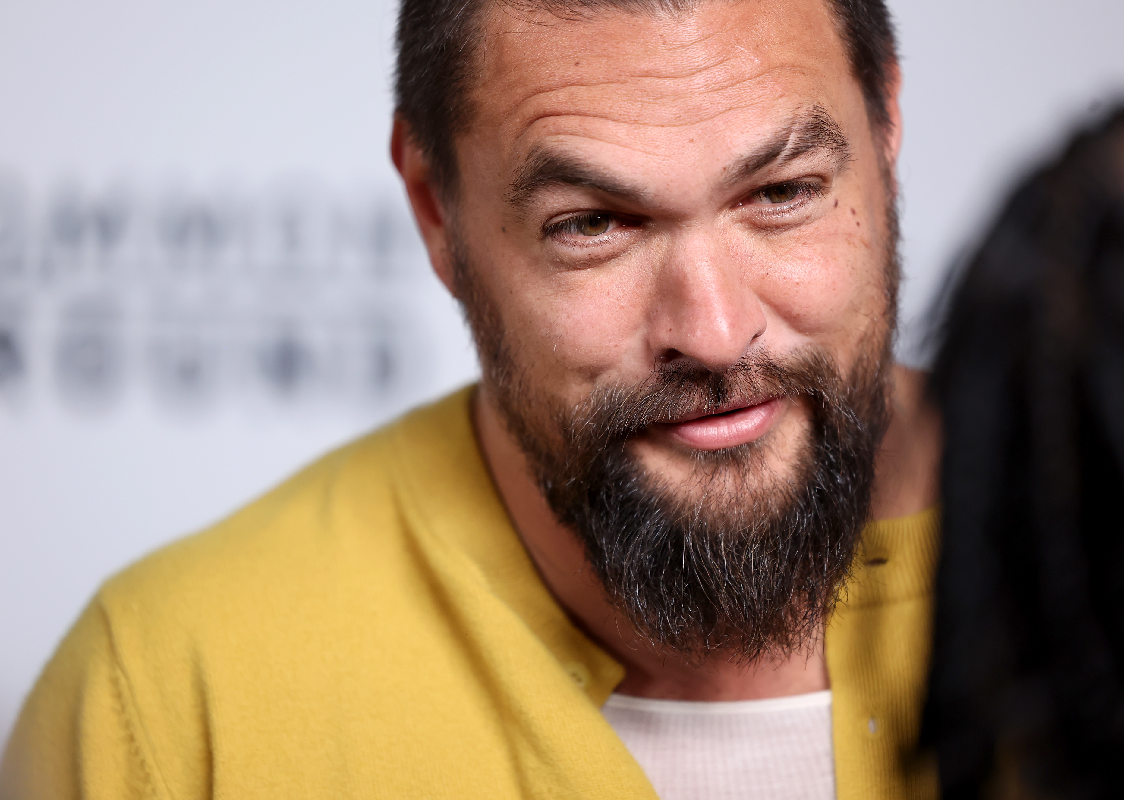 Jason Momoa in a casual shirt smiles while speaking to an unidentified person at an event
