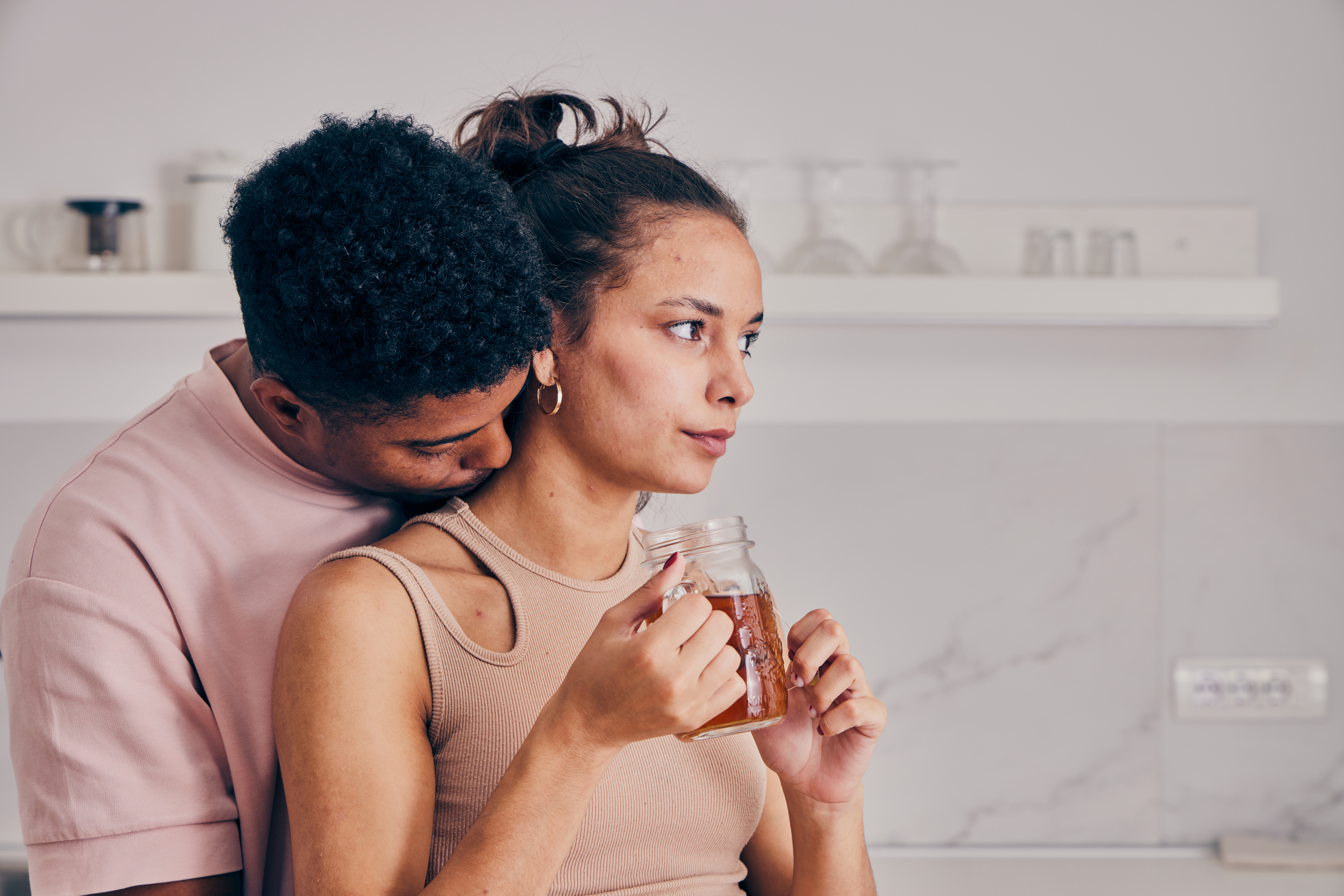 A man hugs a woman from behind as she holds a mason jar with a drink. Both appear thoughtful and are standing in a kitchen
