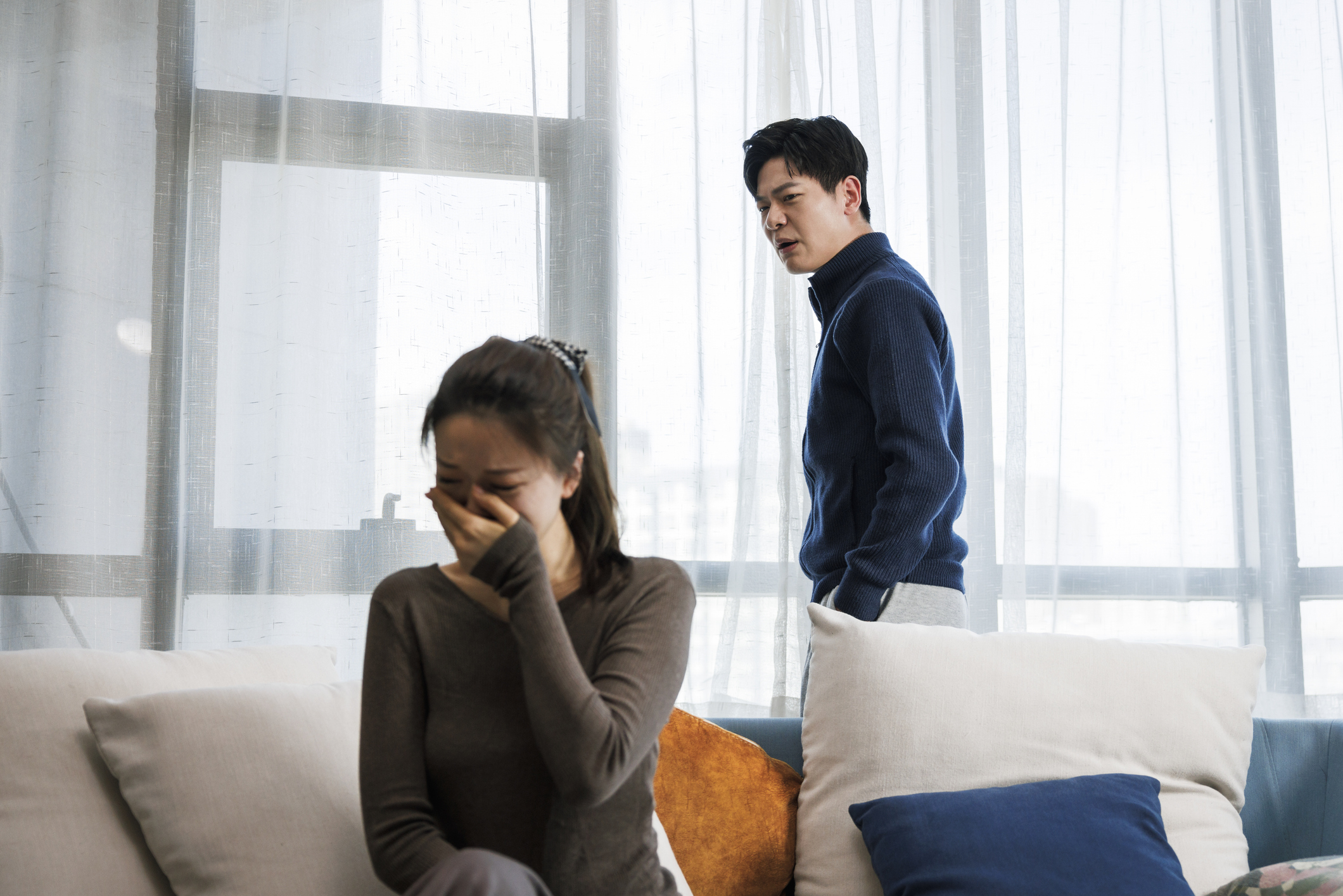 Man in a blue sweater stands with a distressed expression behind a crying woman on a sofa, whose face is in her hands