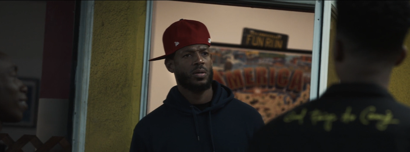 Marlon Wayans in a dark hoodie and red cap standing indoors, engaging in a conversation