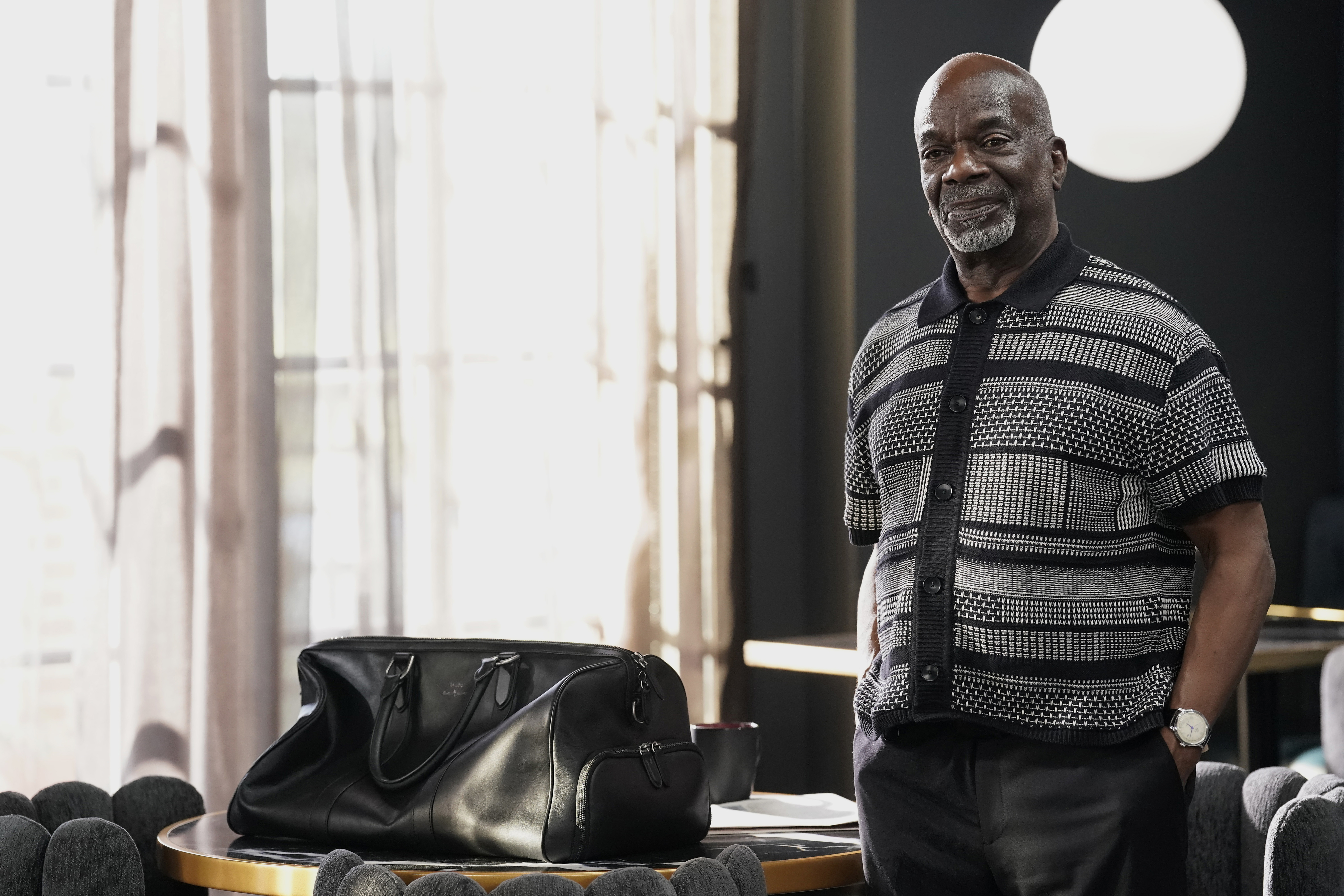 Geoffrey stands by a table with a black duffel bag, wearing a patterned shirt and smiling in a relaxed setting