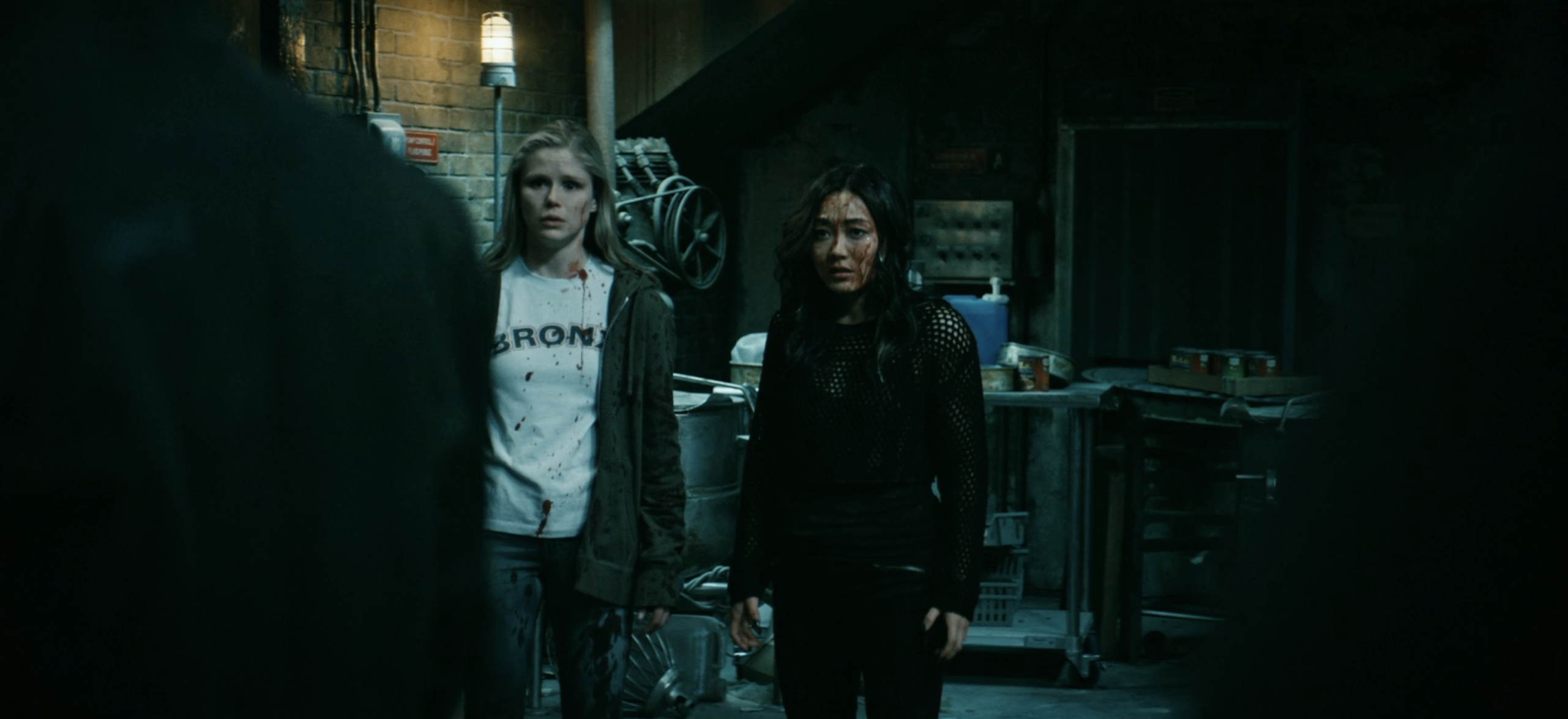 Two women, one in a white shirt and jacket standing with another in a black outfit, appear tense in a dimly lit industrial setting