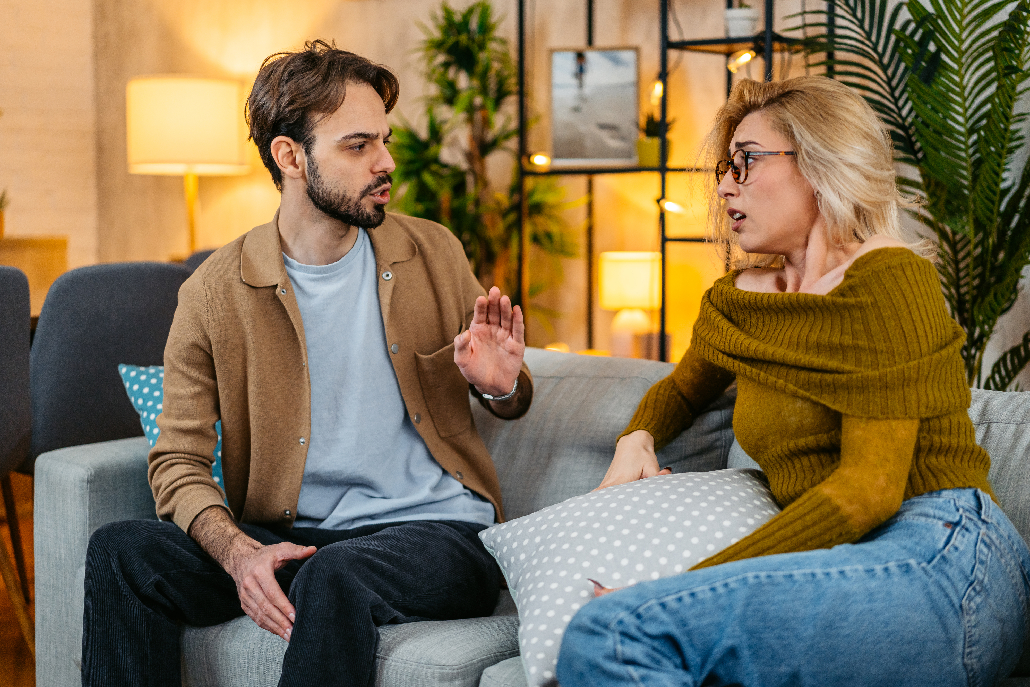 A man and a woman sit on a couch engaged in an intense conversation. The man is gesturing with his hand, while the woman looks defensive