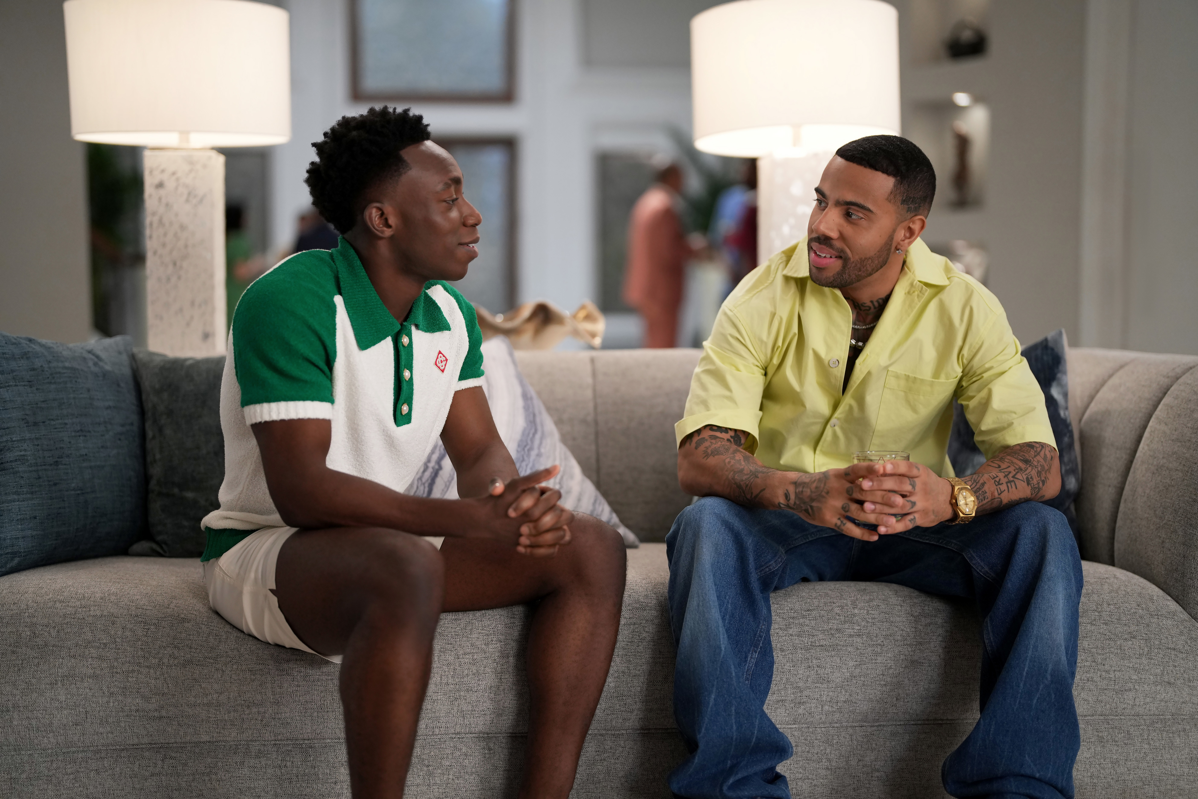 Sam Adegoke in a casual green and white polo shirt, and RJ Cyler in a yellow button-up shirt with jeans, sit talking on a couch in a modern living room setup
