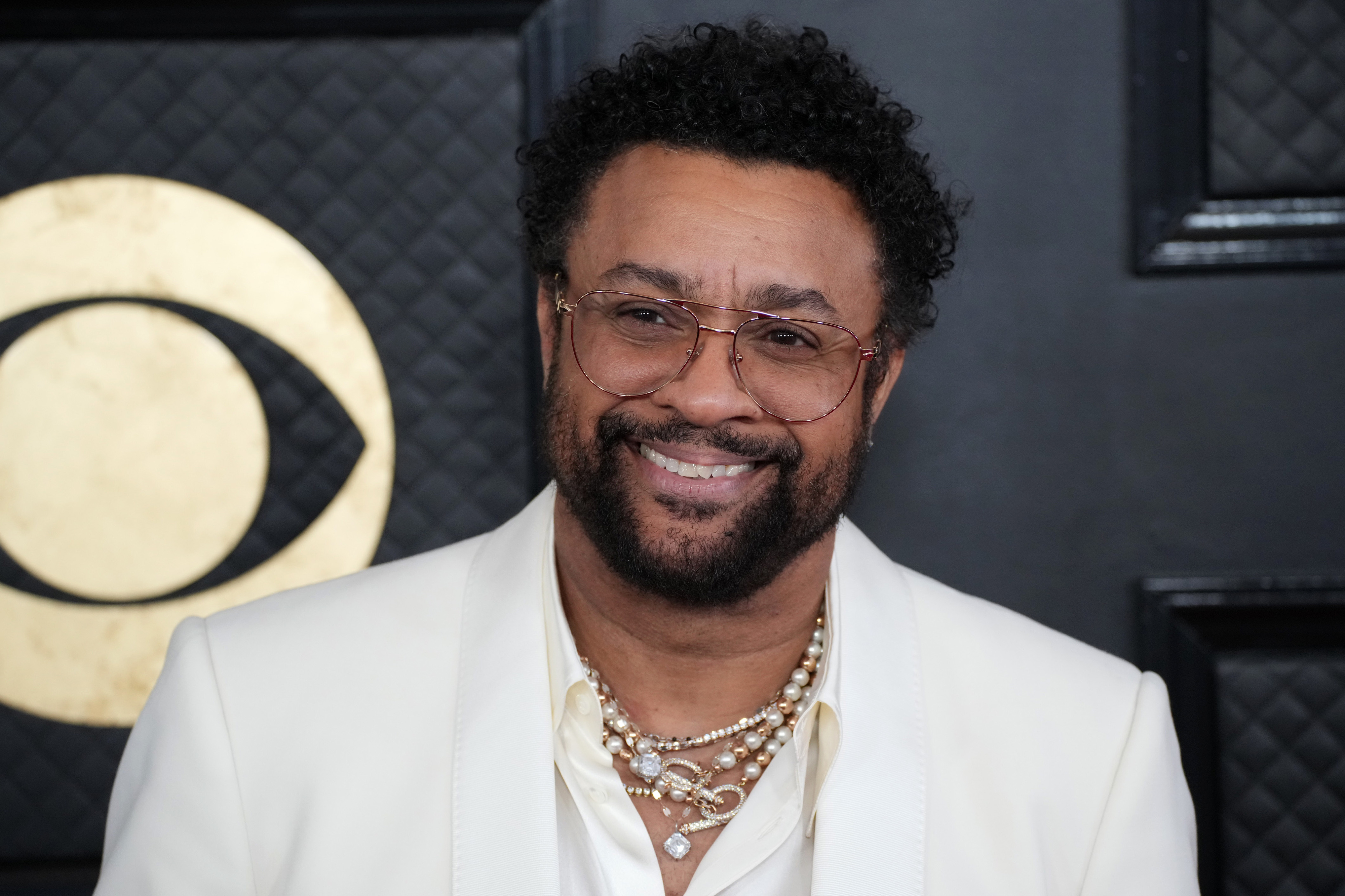 Shaggy wears a white suit with layered necklaces and glasses, smiling against a backdrop at a celebrity event