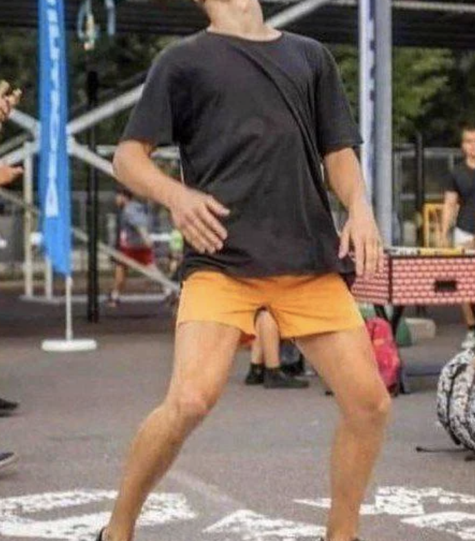 Person in an outdoor setting wearing a black t-shirt, orange shorts, and black shoes, bent backward while balancing on one leg
