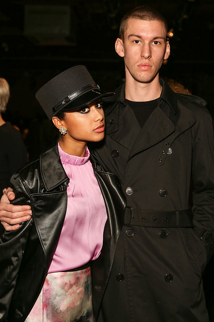 Two individuals posing for a photo. The woman is wearing a leather jacket, a pink top, a floral skirt, and a stylish hat. The man is in a black trench coat. Names unknown