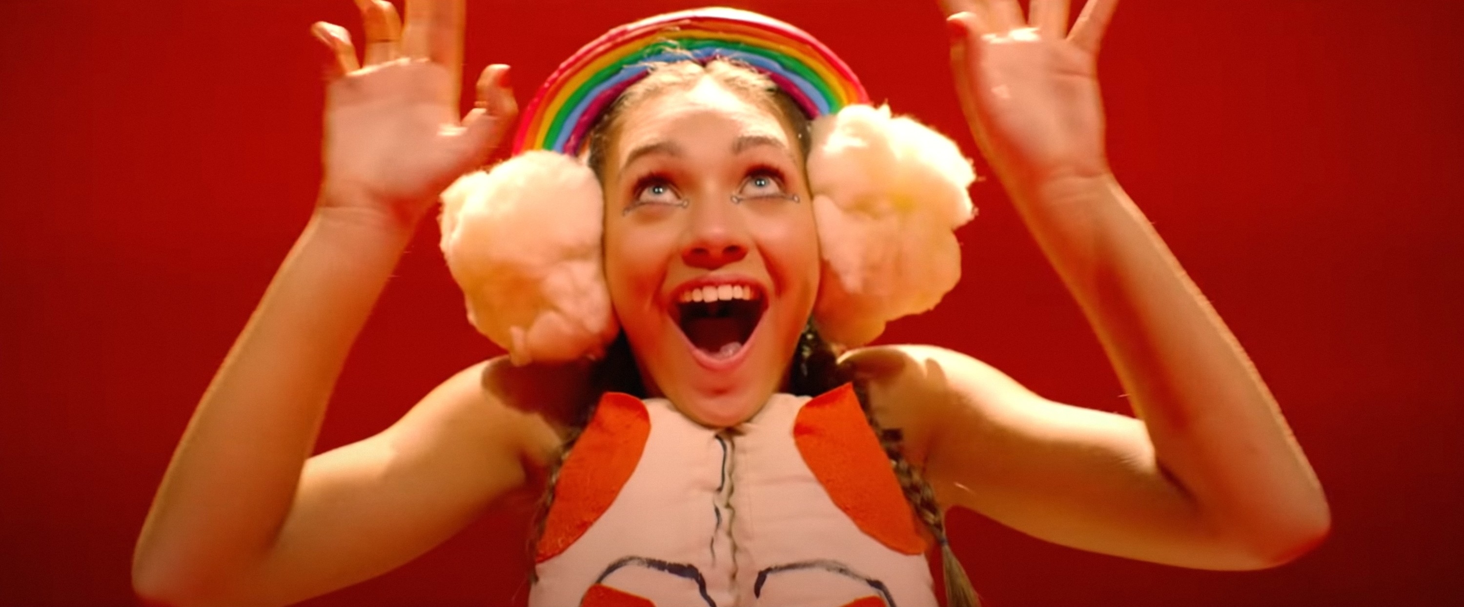 A person wearing a rainbow hat, large pink earmuffs, and a colorful outfit is excitedly raising their hands against a red background