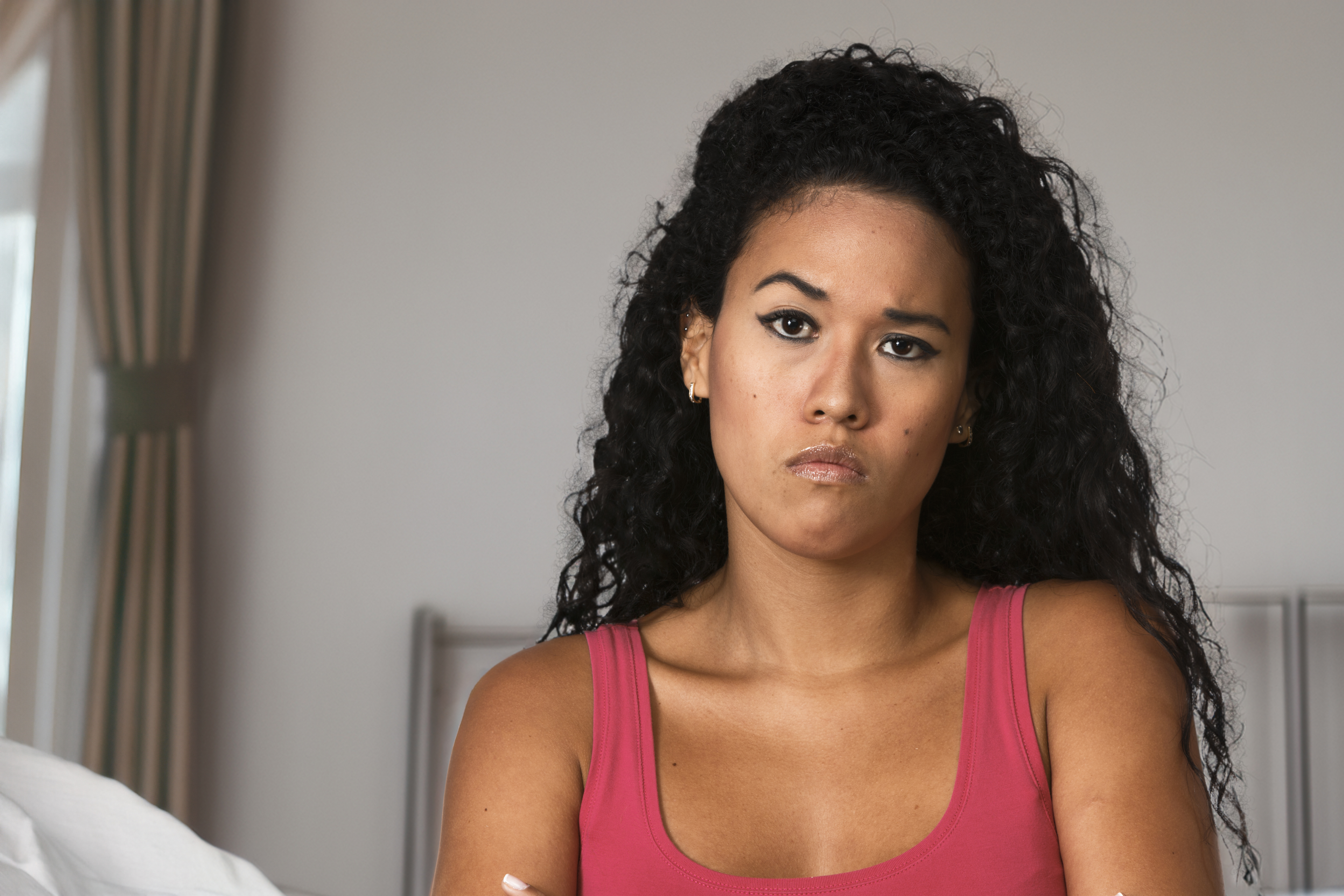 A woman with long curly hair wearing a sleeveless top looks upset or frustrated, with arms crossed. She is indoors with a window and curtains visible in the background