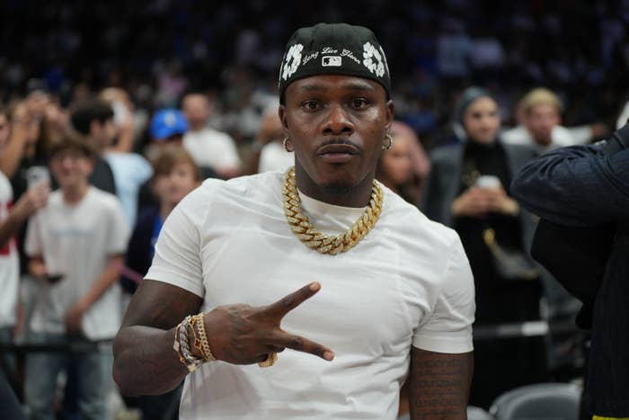 DaBaby wearing a white shirt and gold chains, making a peace sign gesture at a public event