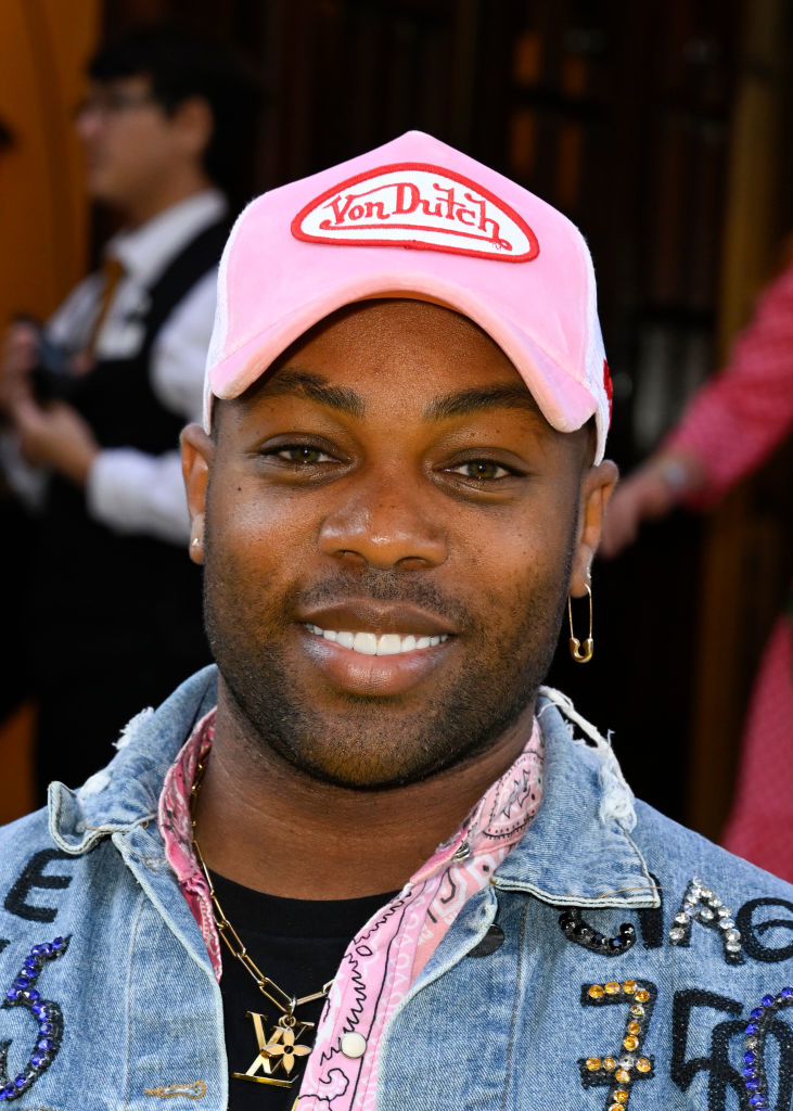 Todrick Hall wears a Von Dutch cap, denim jacket with colorful patches, pink bandana, and layered necklaces at a public event