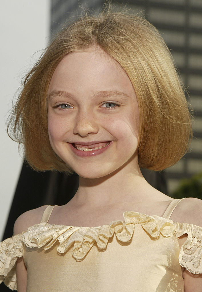 A young girl, Dakota Fanning, smiles brightly with a shoulder-length bob haircut and wearing a ruffled top, standing outside with a building in the background