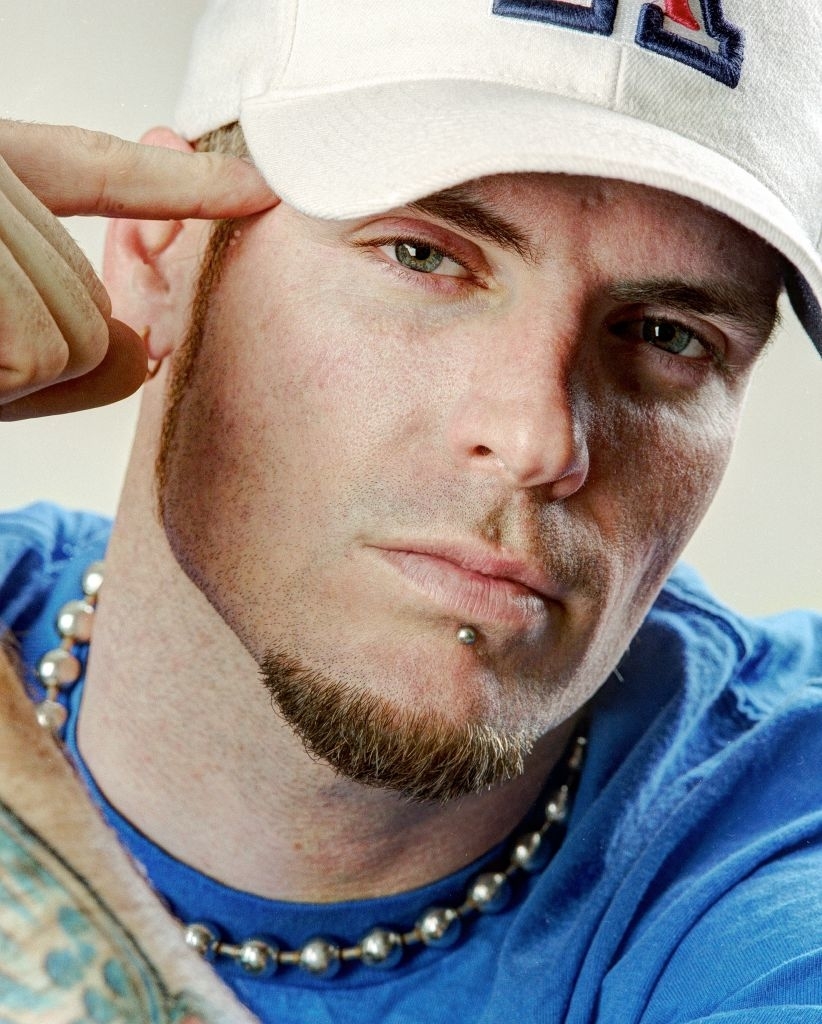 Vanilla Ice in a close-up shot, pointing to his temple, wearing a white cap, blue t-shirt, beaded necklace, and sporting a goatee and lip piercing