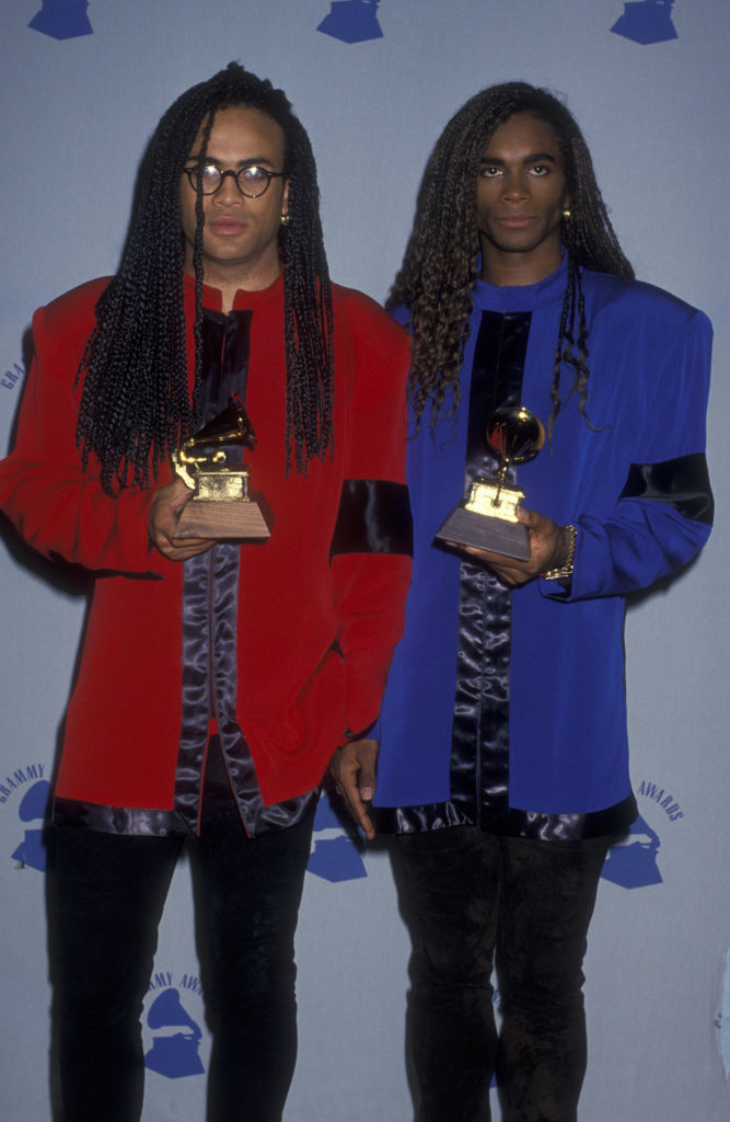 Two people holding Grammy Awards, both with long braided hair and wearing oversized, shiny jackets, one in red and the other in blue, with black accents