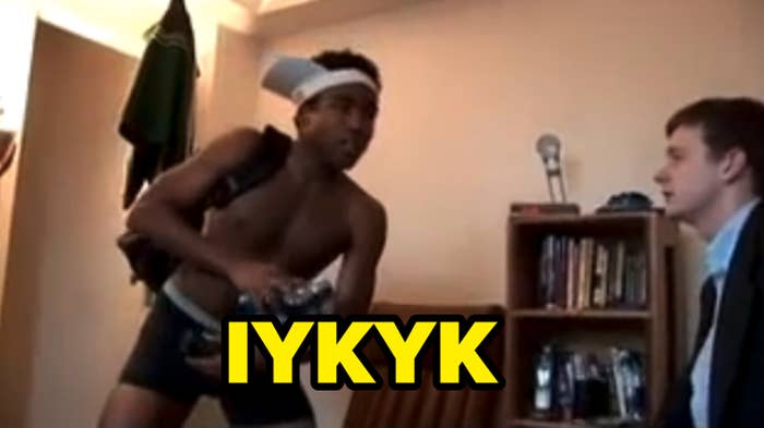 Donald Glover in shorts, backpack, and headband holding water bottles, talking to a seated person in suit by bookshelf