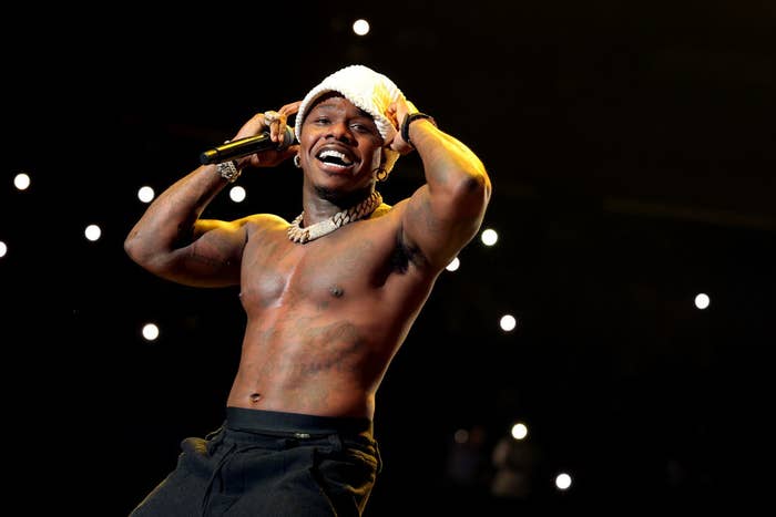 DaBaby, shirtless and wearing a bucket hat, performs on stage holding a microphone. Background shows lit-up audience