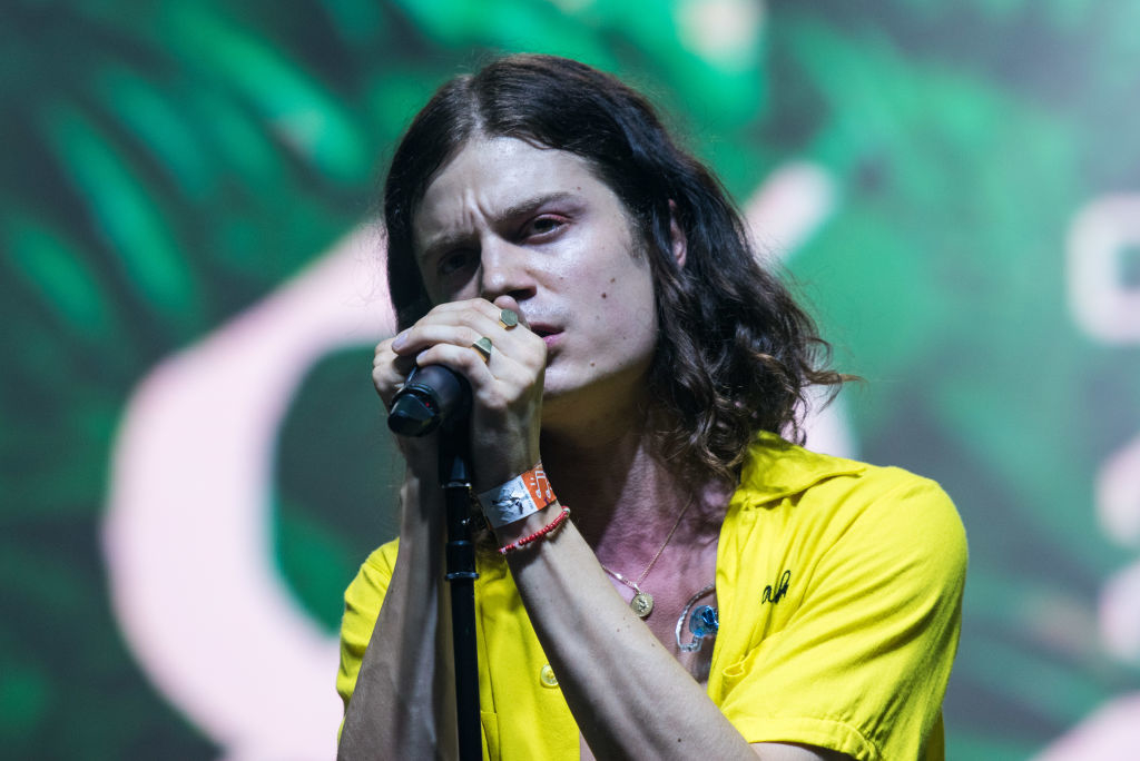 Long-haired singer performing on stage, wearing a yellow shirt and holding a microphone close to his mouth