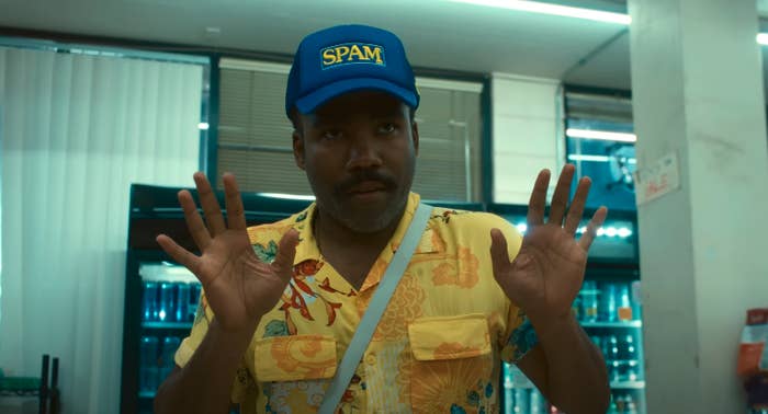 Donald Glover in a store, wearing a hat with the word "SPAM" on it, and a Hawaiian shirt, holds up his hands in front of him