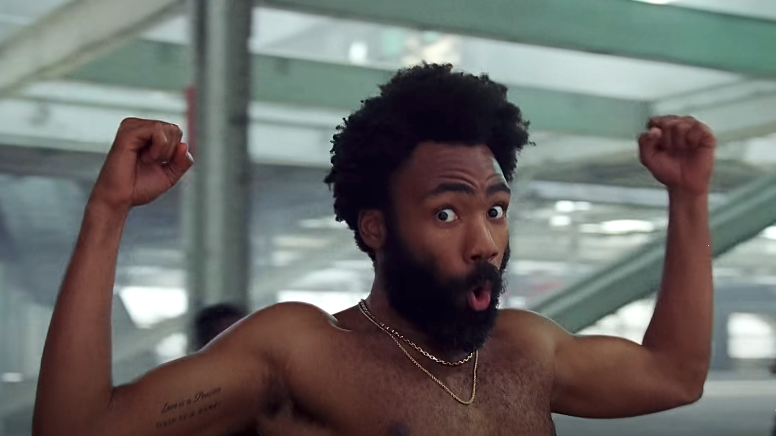 Donald Glover is posing shirtless with his arms flexed and mouth open in a surprised expression. He is wearing a gold chain necklace