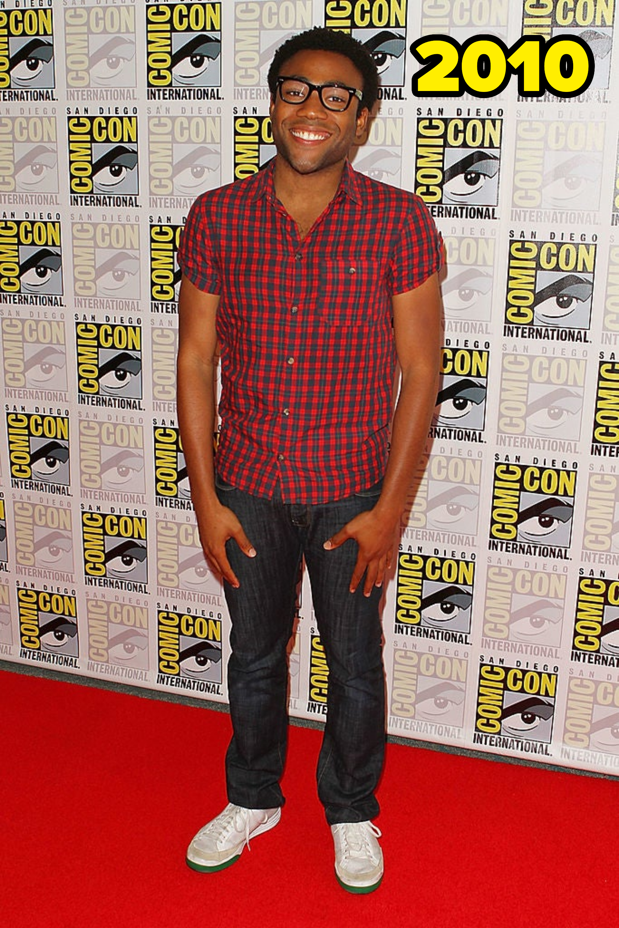 Donald Glover stands on the Comic-Con red carpet wearing a plaid shirt, jeans, and white sneakers. He is smiling in front of the Comic-Con International backdrop