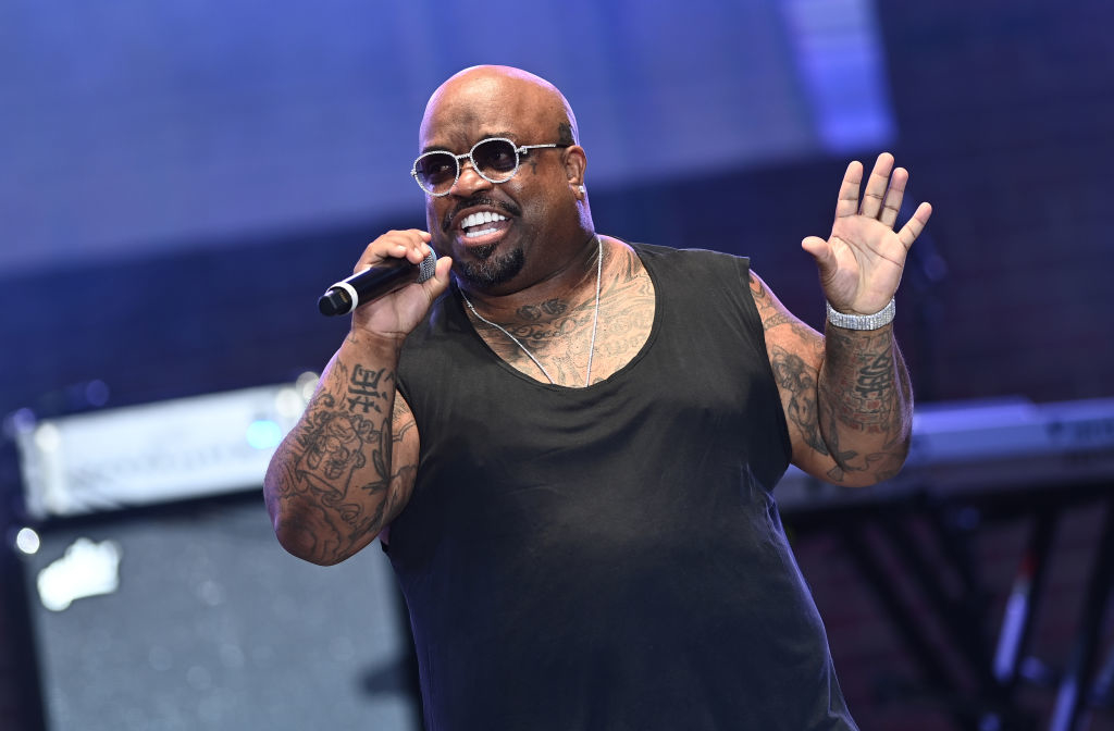 CeeLo Green performing on stage, wearing a black sleeveless top and round sunglasses, holding a microphone and gesturing with his hand