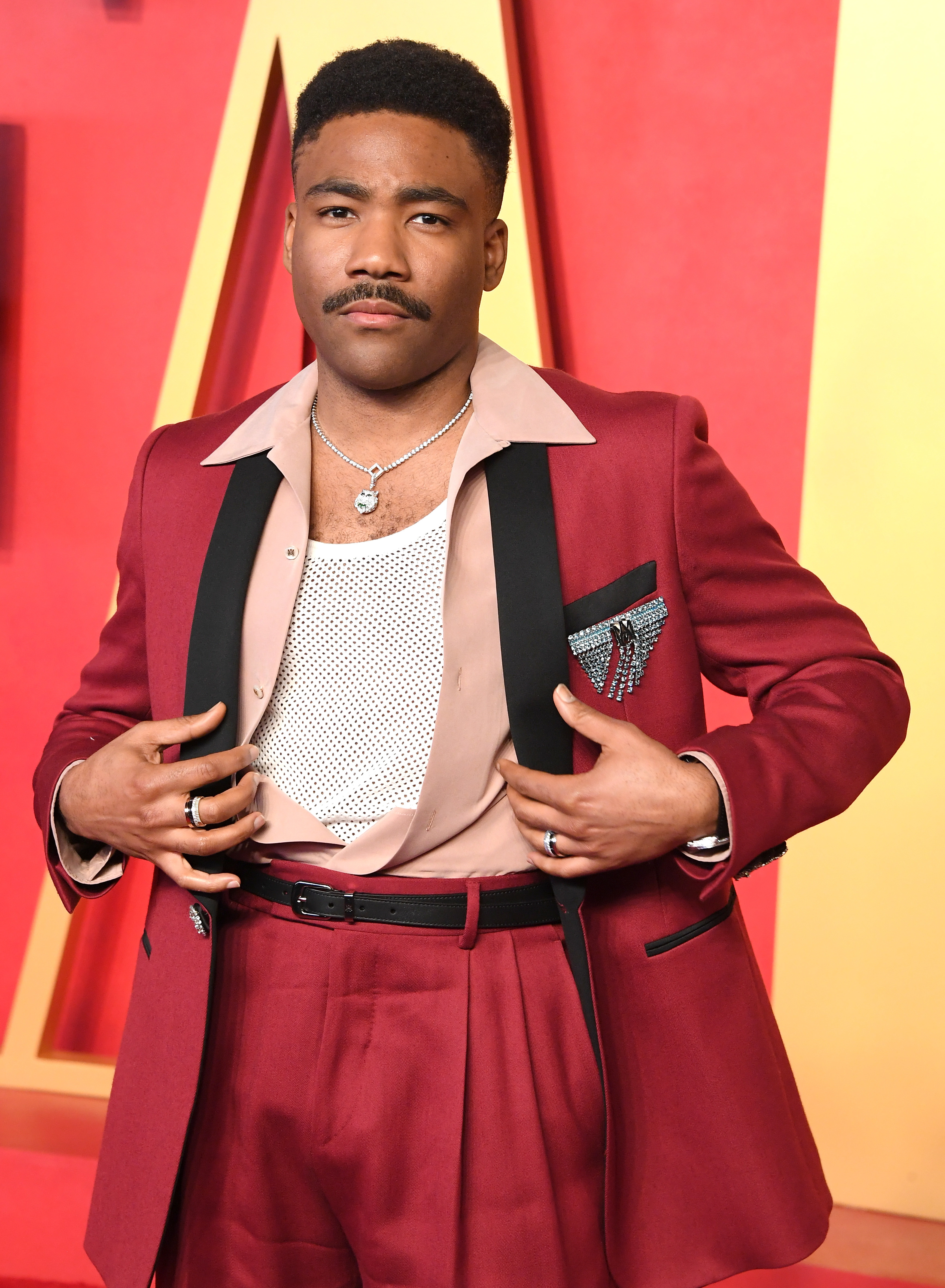 A person poses on the red carpet wearing a stylish red suit with a beige shirt and patterned pocket square