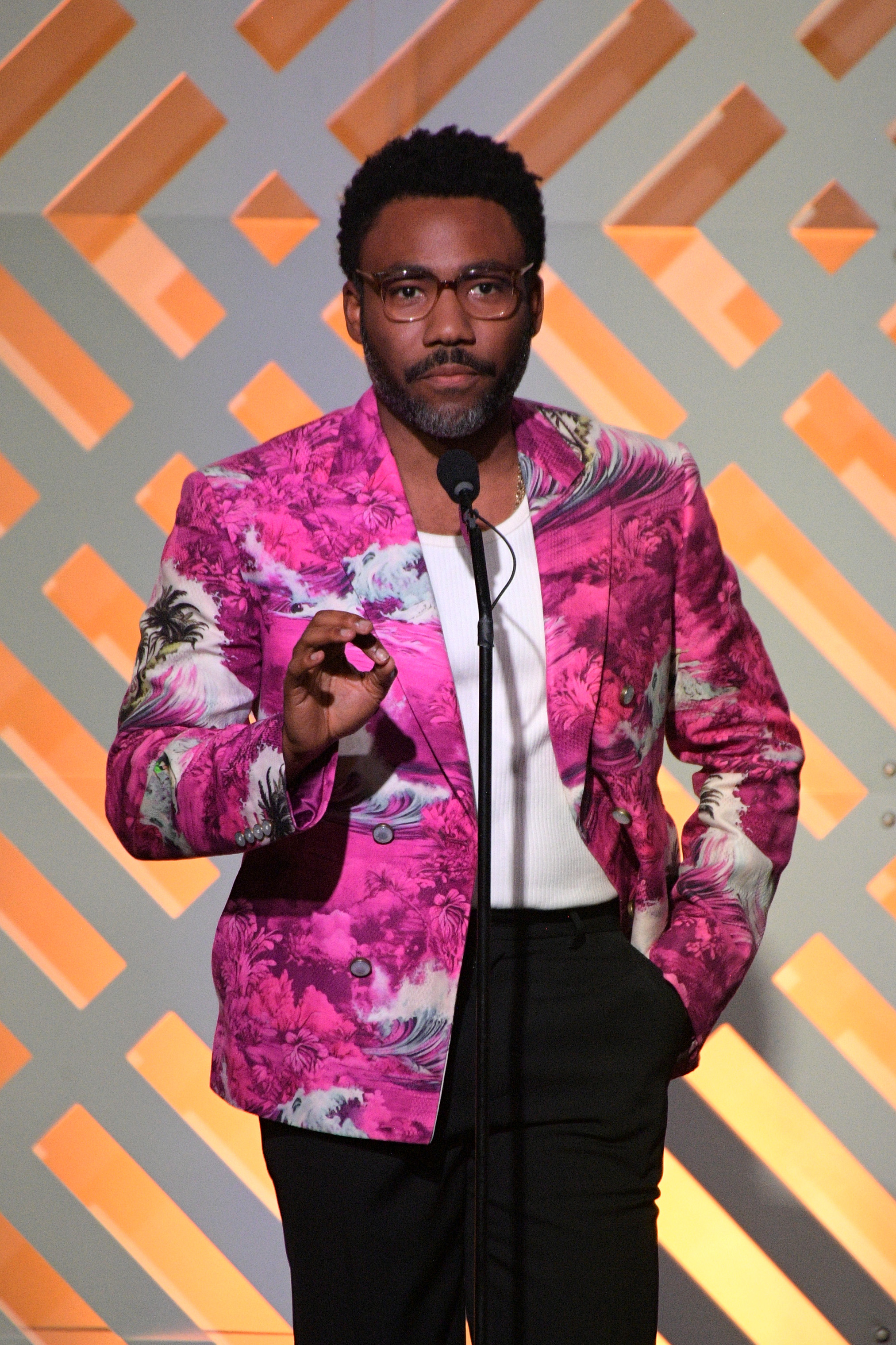 Donald Glover stands at a podium in a pink floral blazer, white shirt, and black pants, speaking at an event