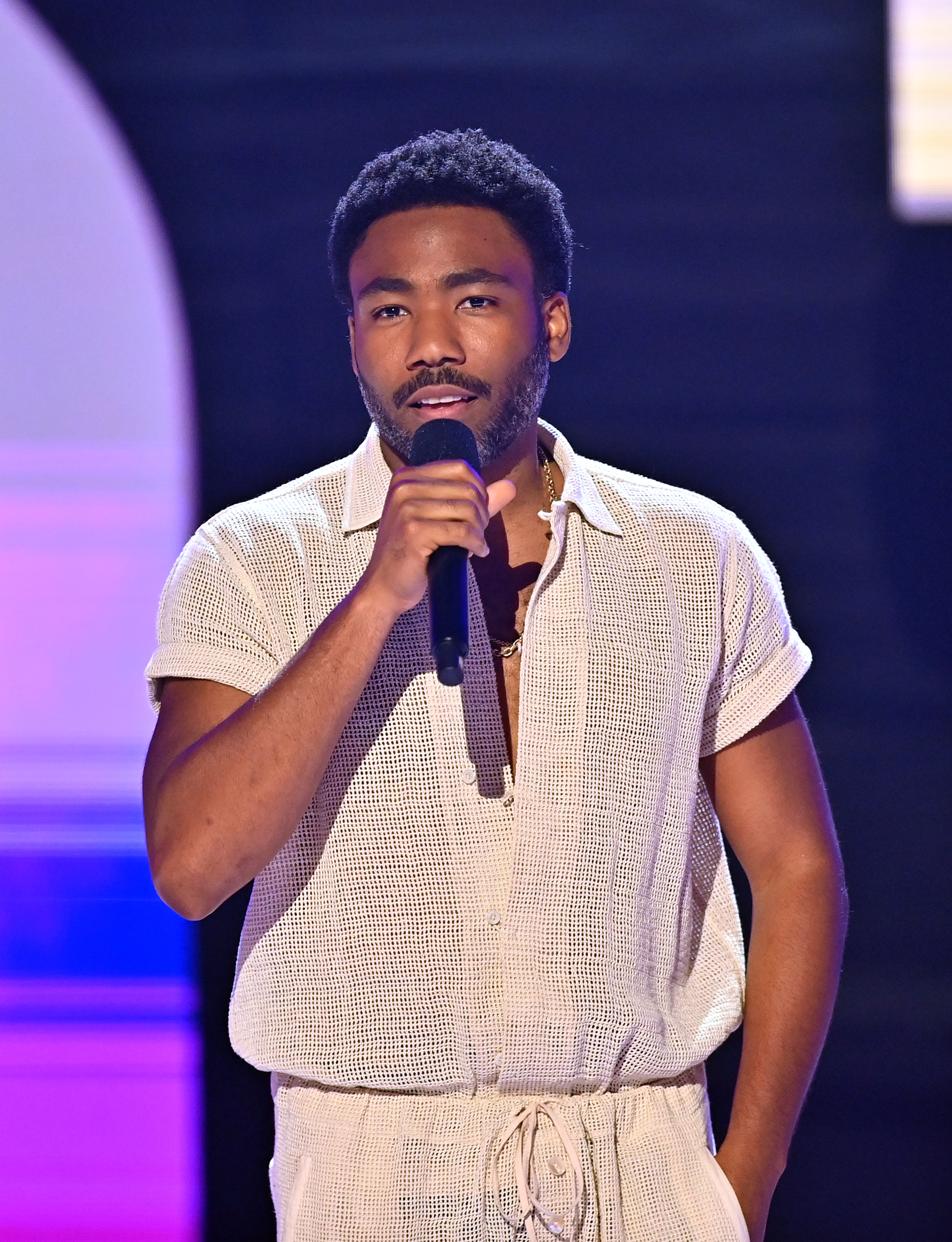 Donald Glover speaks into a microphone while wearing a casual, textured outfit on stage