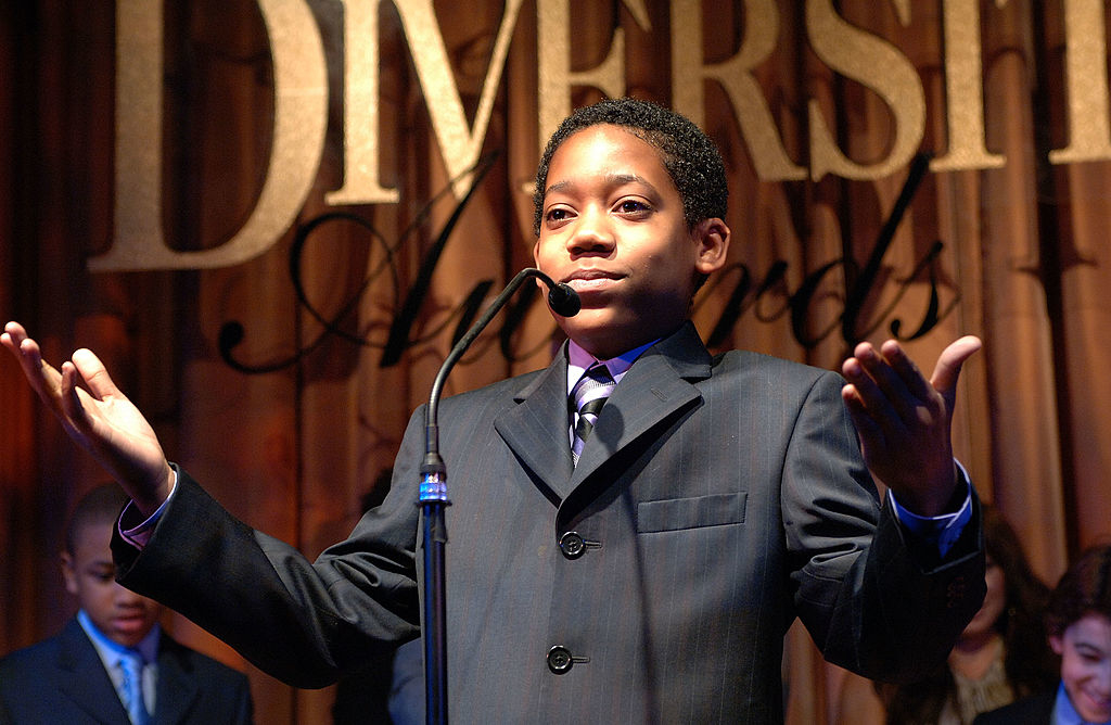 Young person in a suit speaking into a microphone at a formal event with the words "Diverse Awards" in the background