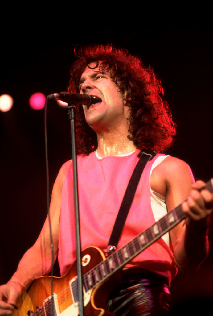 A person with shoulder-length hair plays an electric guitar and sings into a microphone on stage, wearing a sleeveless top
