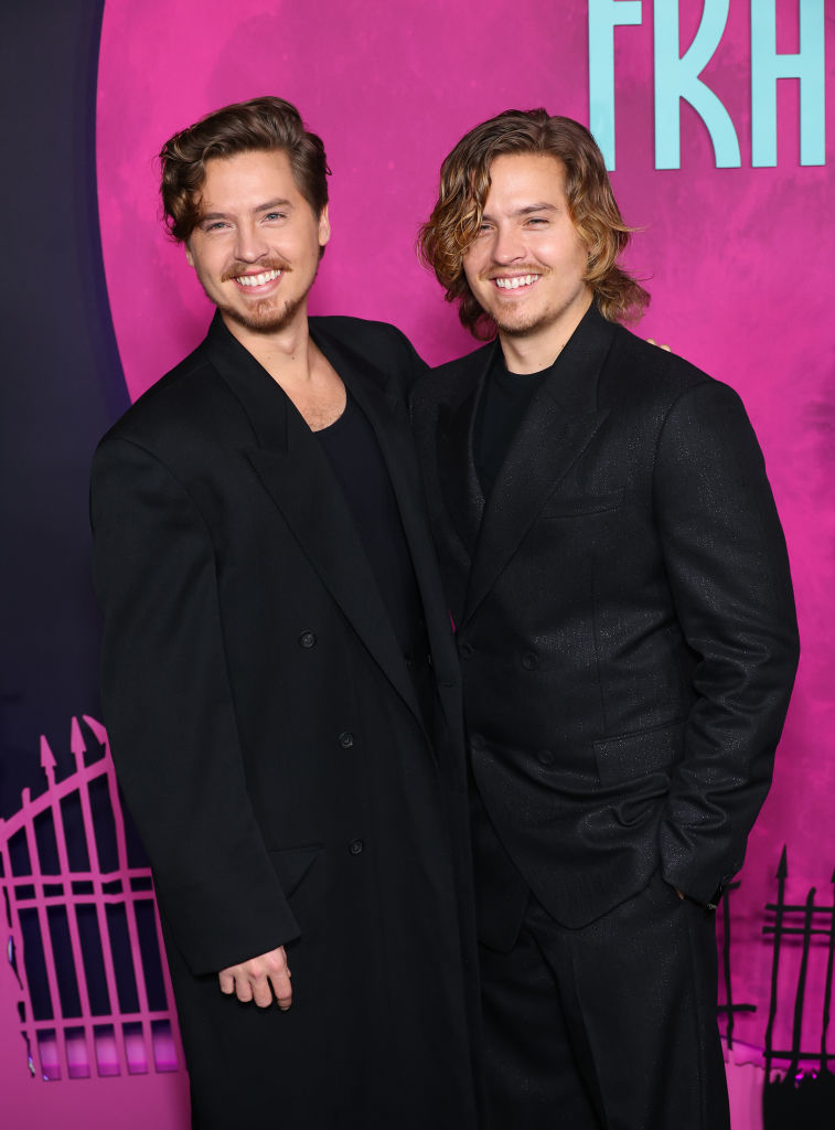 Dylan and Cole Sprouse smile while standing together on a red carpet. Both are wearing dark, stylish suits.